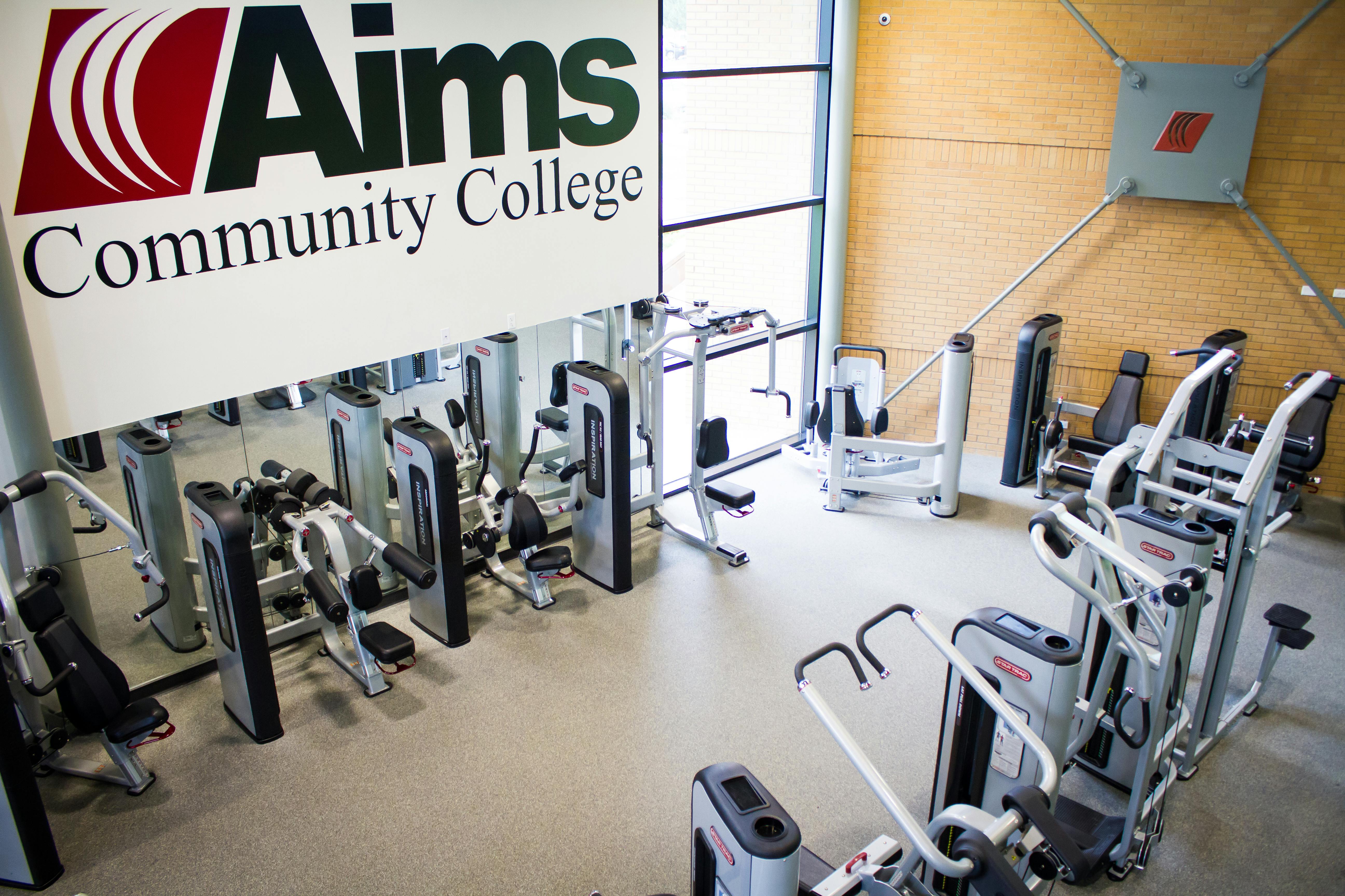 Exercise equipment in rows around edge of room with Aims Community College logo sign hanging above one row