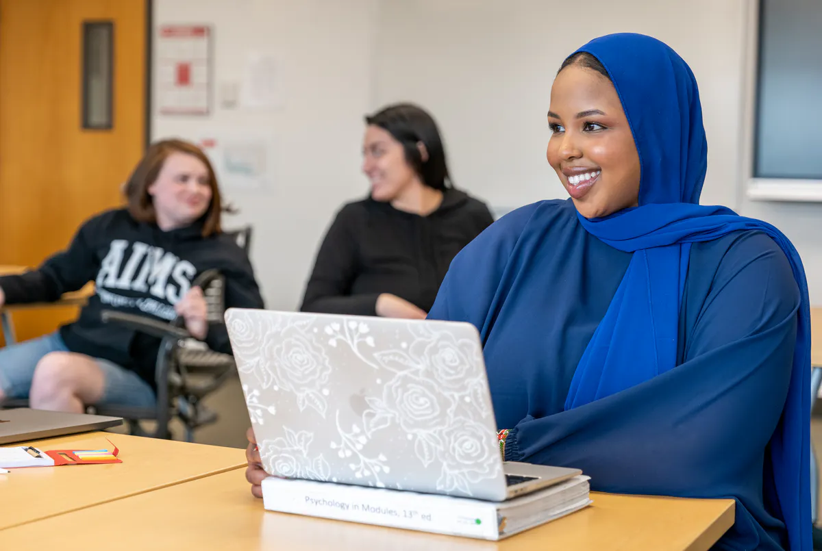 A student sits at a table with a laptop in a classroom
