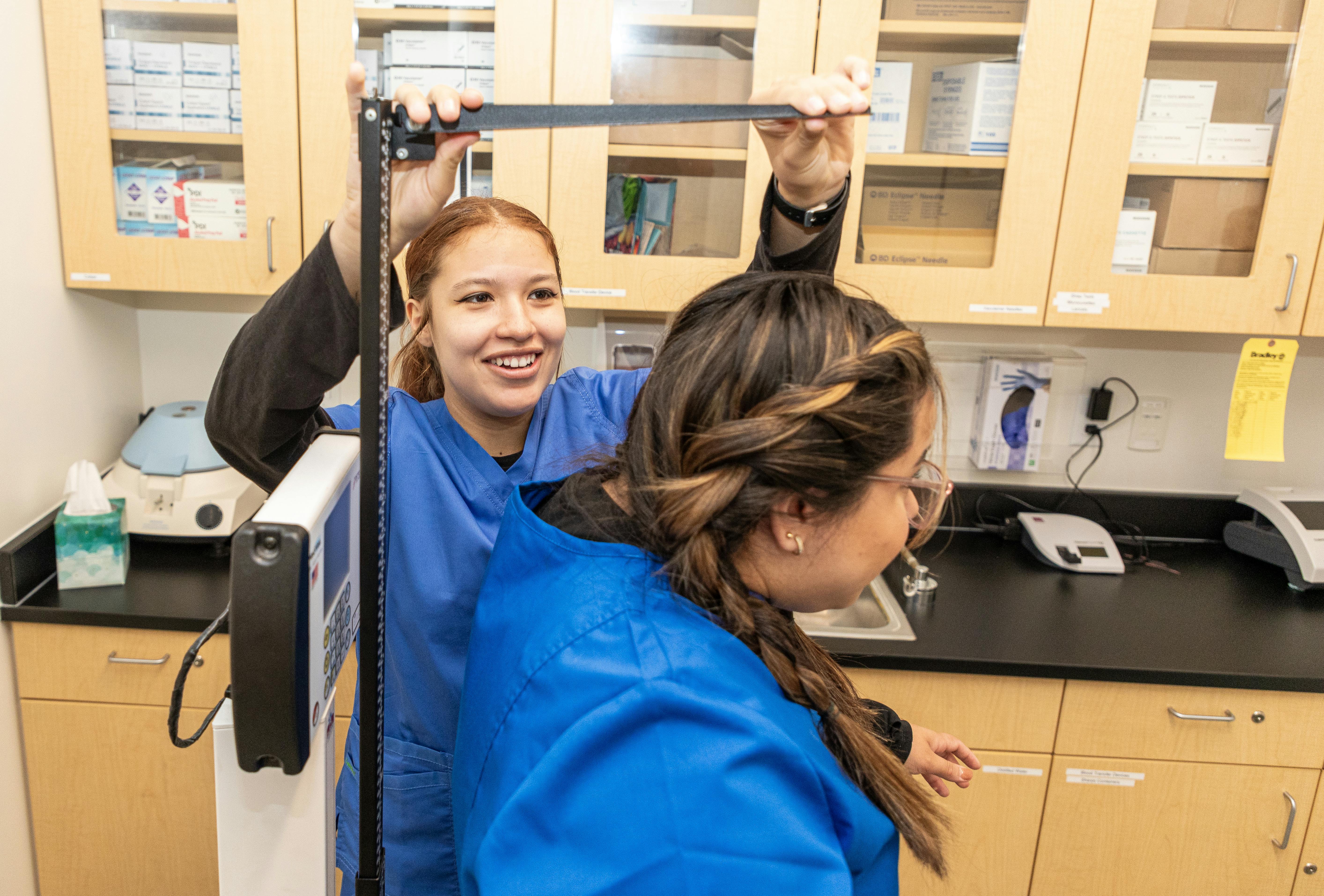 Aims Nurse Aide students wearings scrubs and practicing feeding techniques in a lab setting