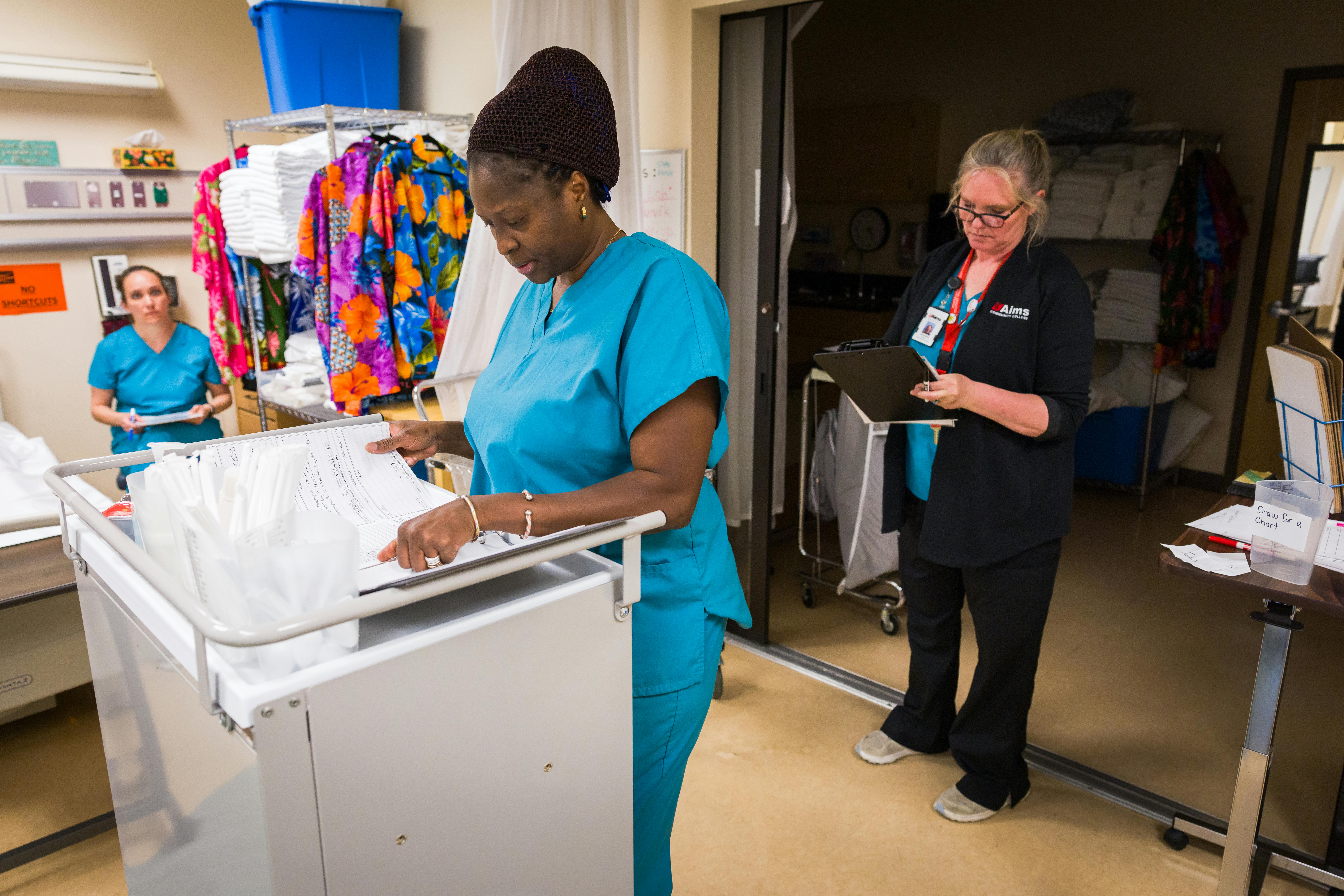 Student wearing blue scrubs standing at cart practicing filling medication orders while instructor and another student look on.