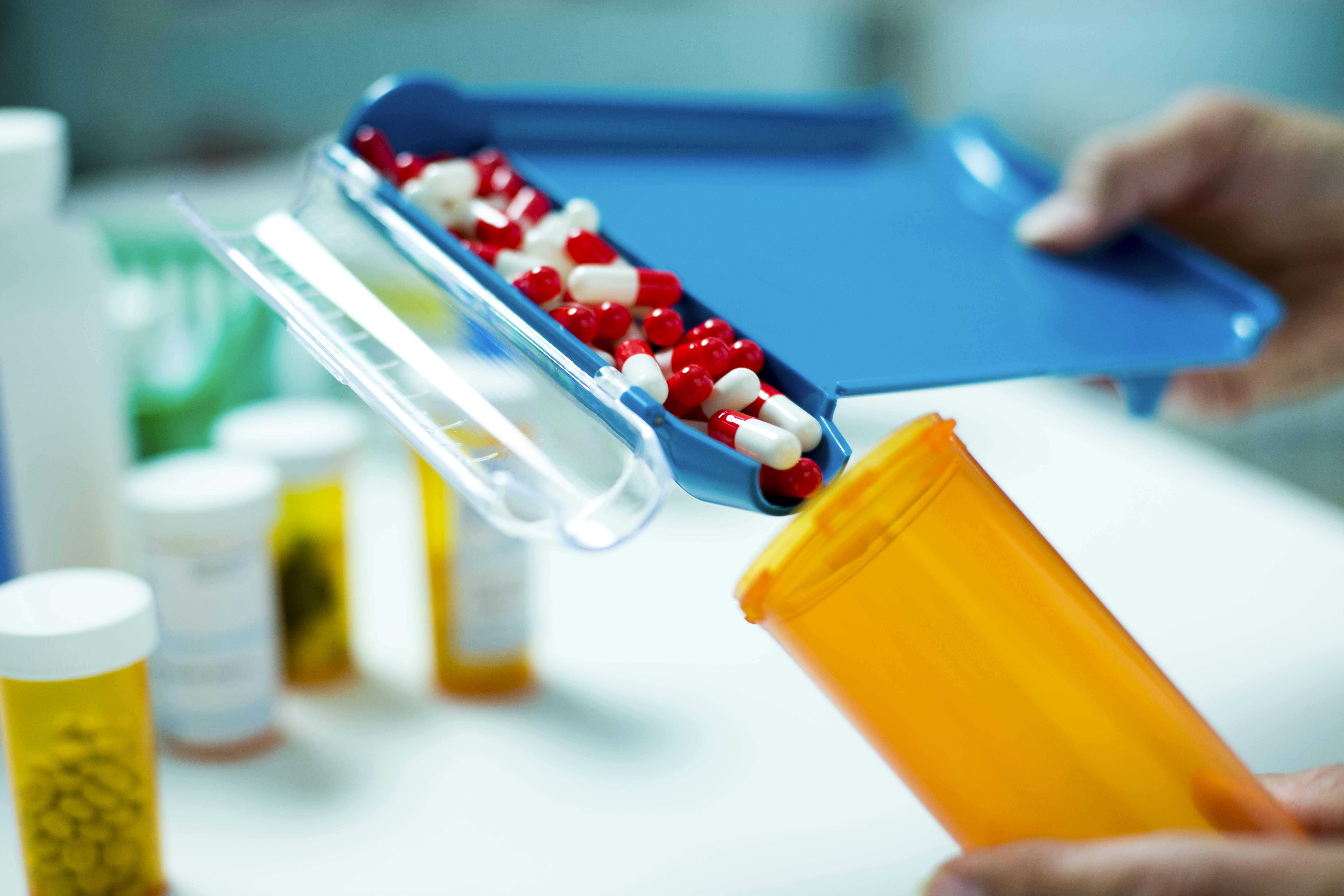 hands holding empty pill bottle using blue dispenser to fill bottle with red and white capsules 