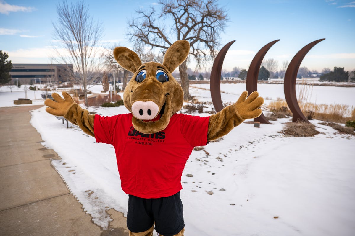 Aim's mascot Arty the Aardvark stands with open arms in front of a snow covered campus