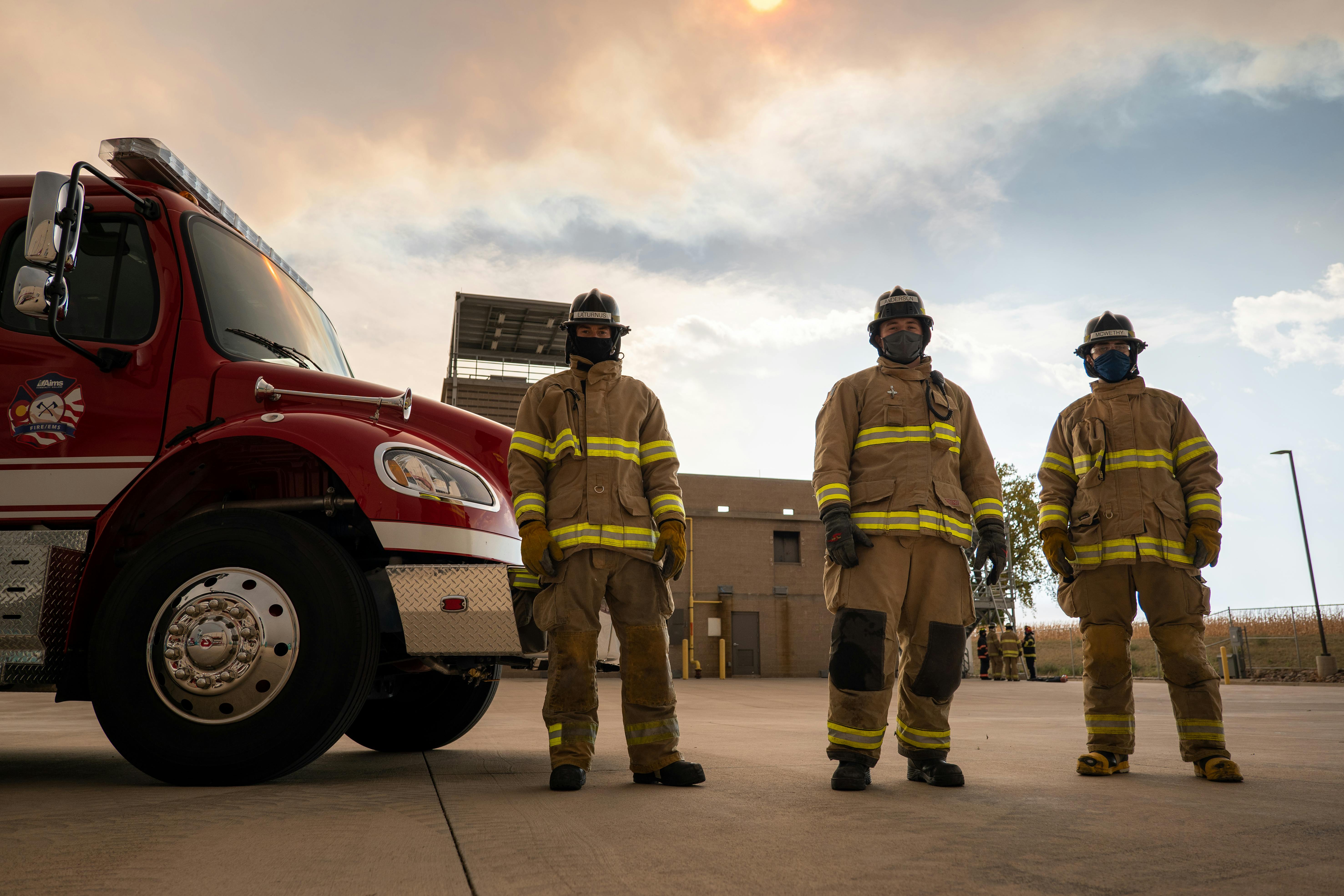Three firefighters stand framed by a sunny sky in front of their firetruck.