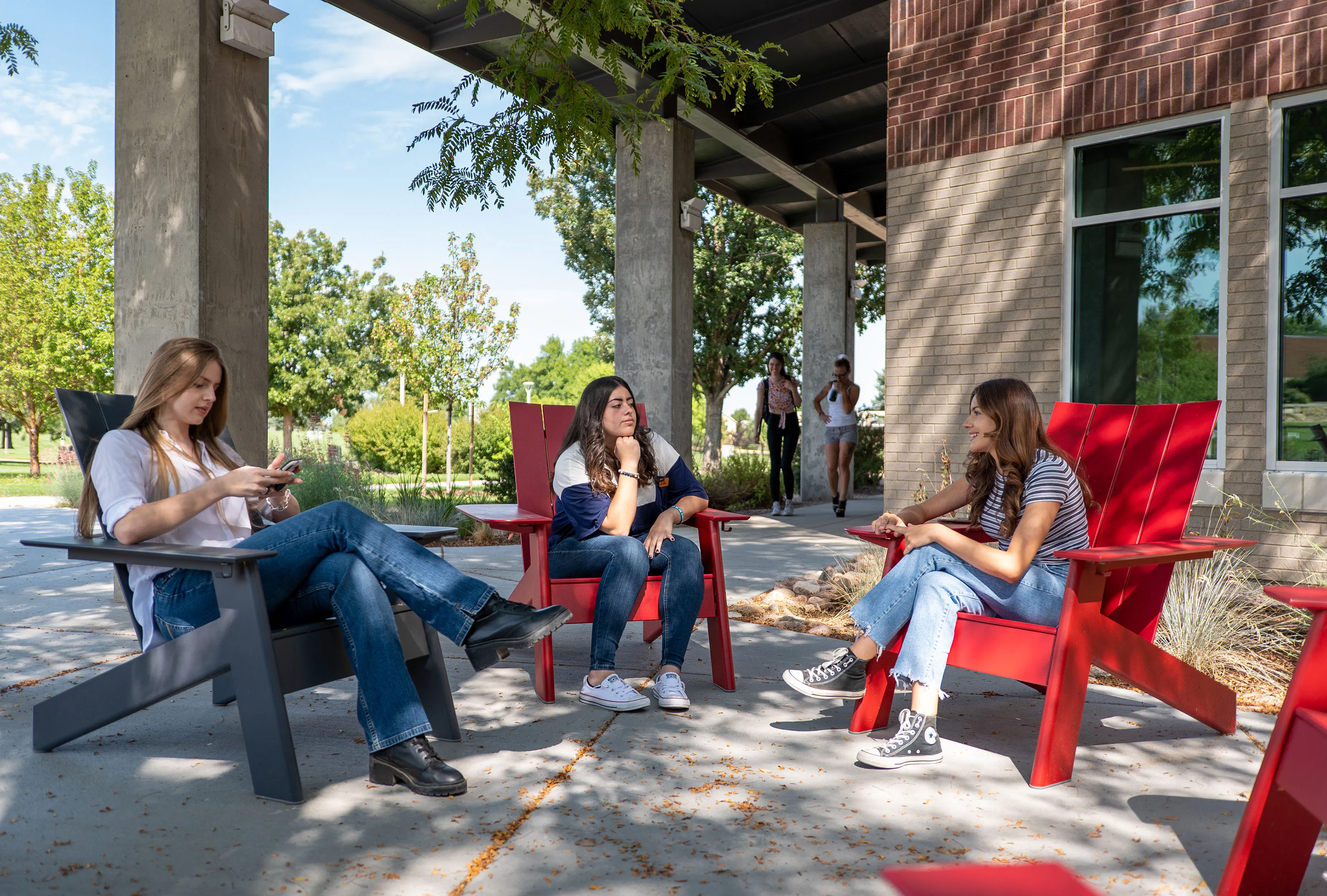 Three students sit outside in red and black Adirondack chairs. They are chatting with each other
