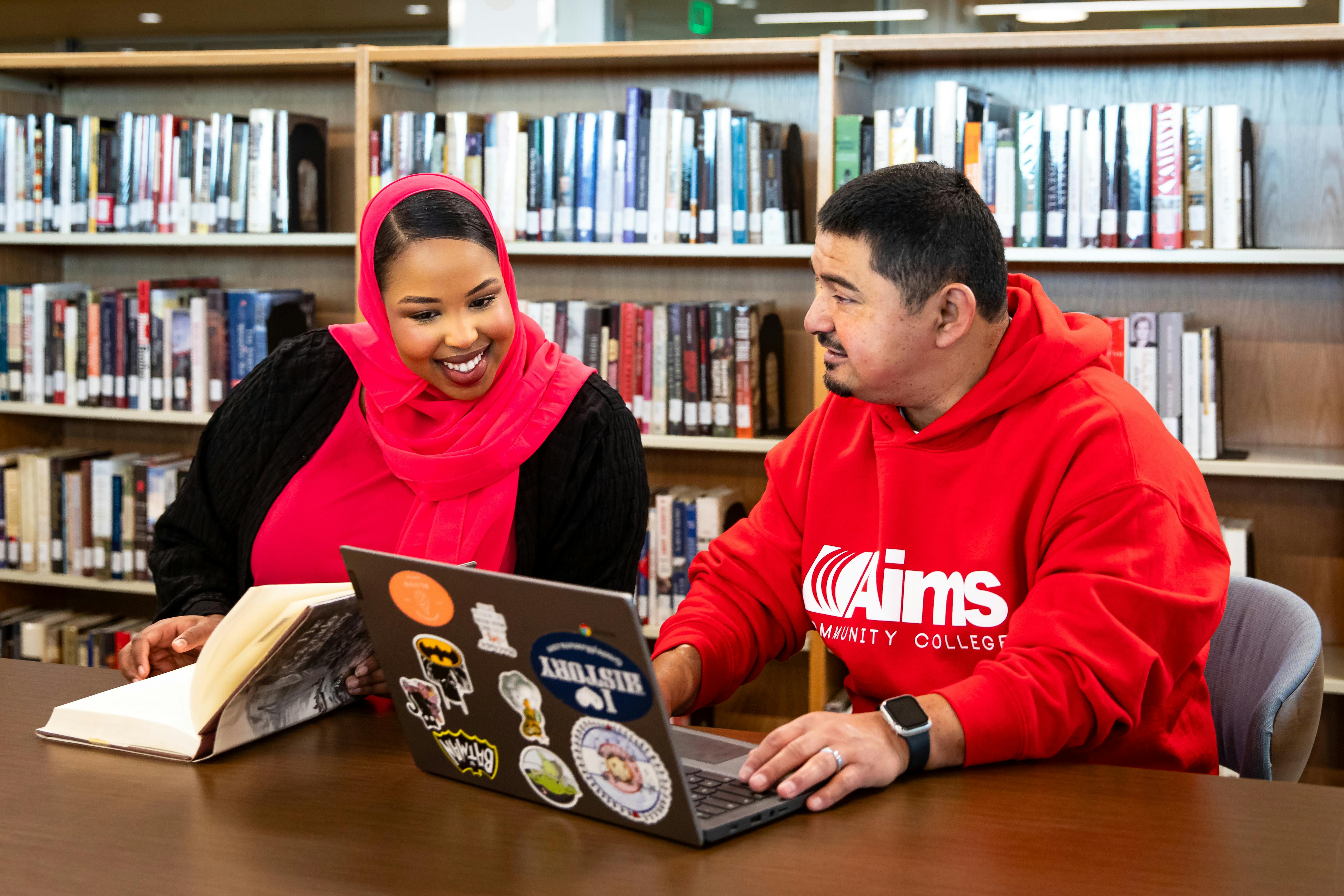 Two students sitting side by side at a table looking at a book and an open laptop