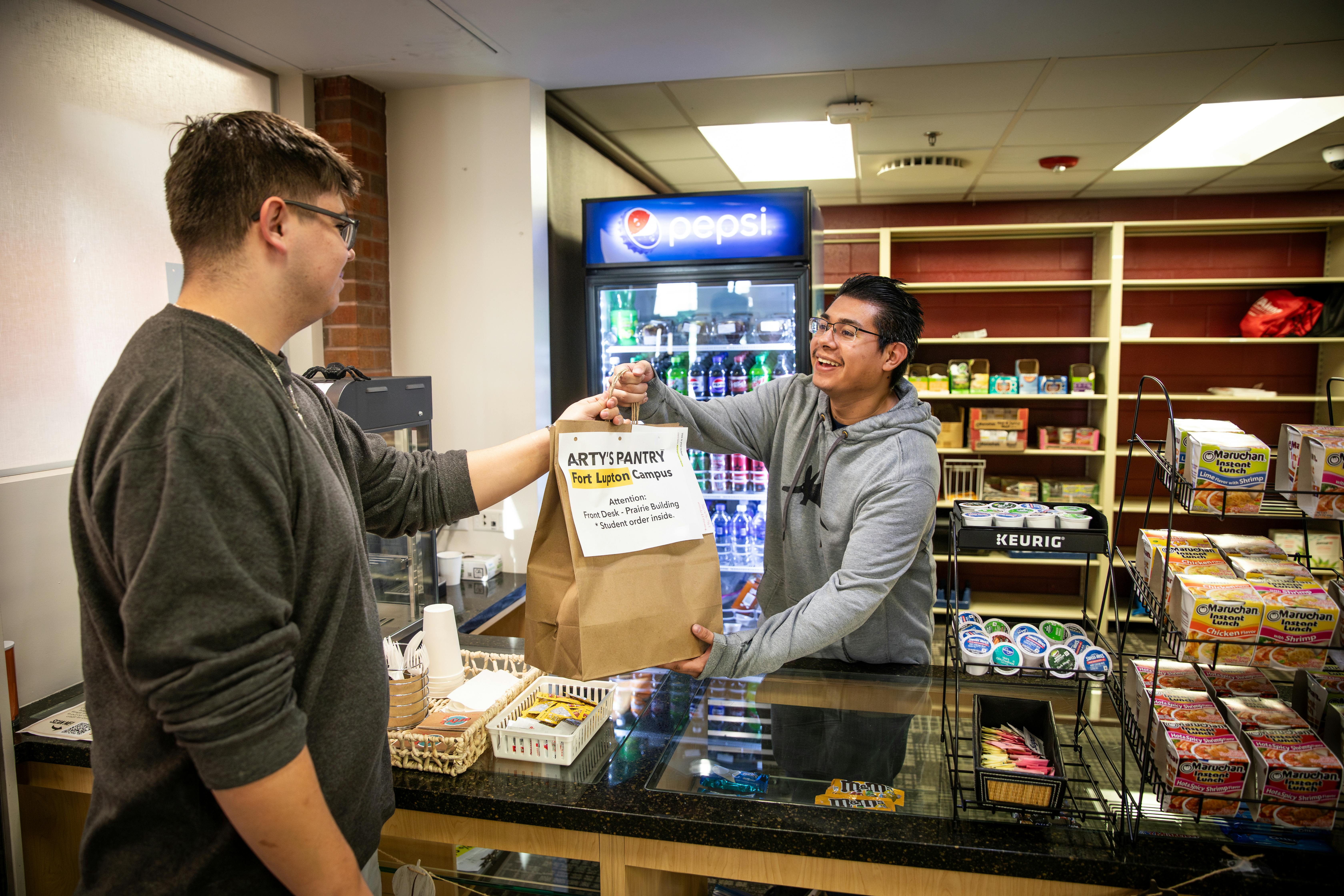 Volunteer at pantry hands paper bag of groceries to another student across a counter, shelves of food behind him