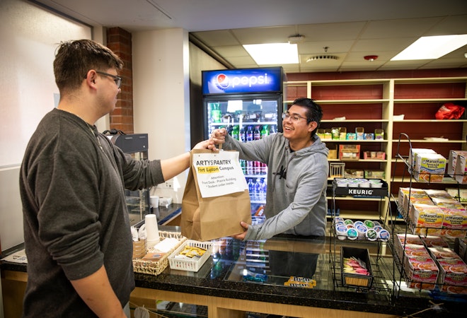 Volunteer at pantry hands paper bag of groceries to another student across a counter, shelves of food behind him