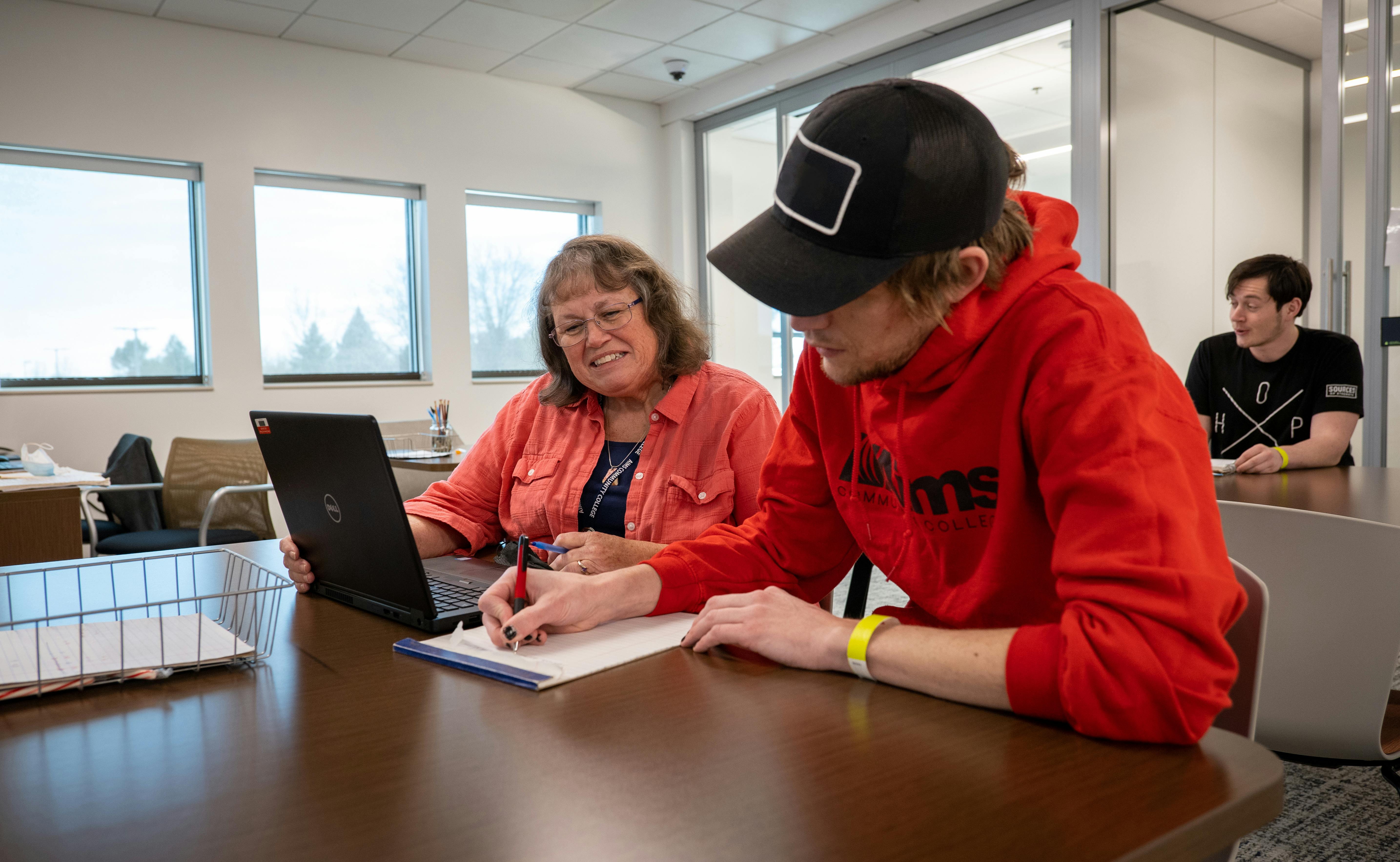 student in red shirt and black ball cap sitting at table while writing on notepad next to woman wearing red shirt and glasses in front of open laptop, smiling at student