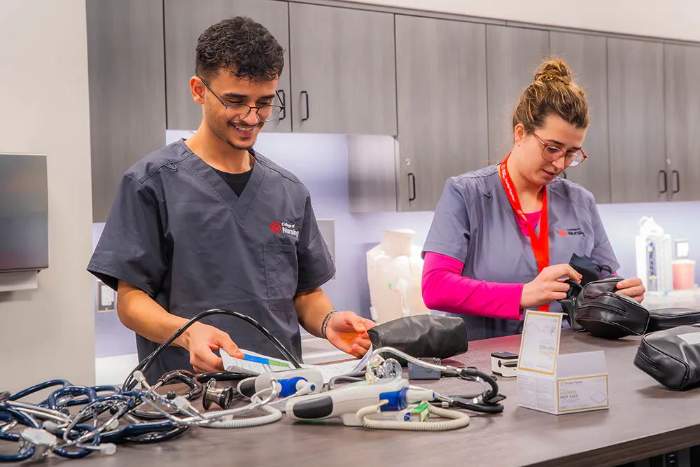 Two students in clinical scrubs handle medical training equipment on a lab counter