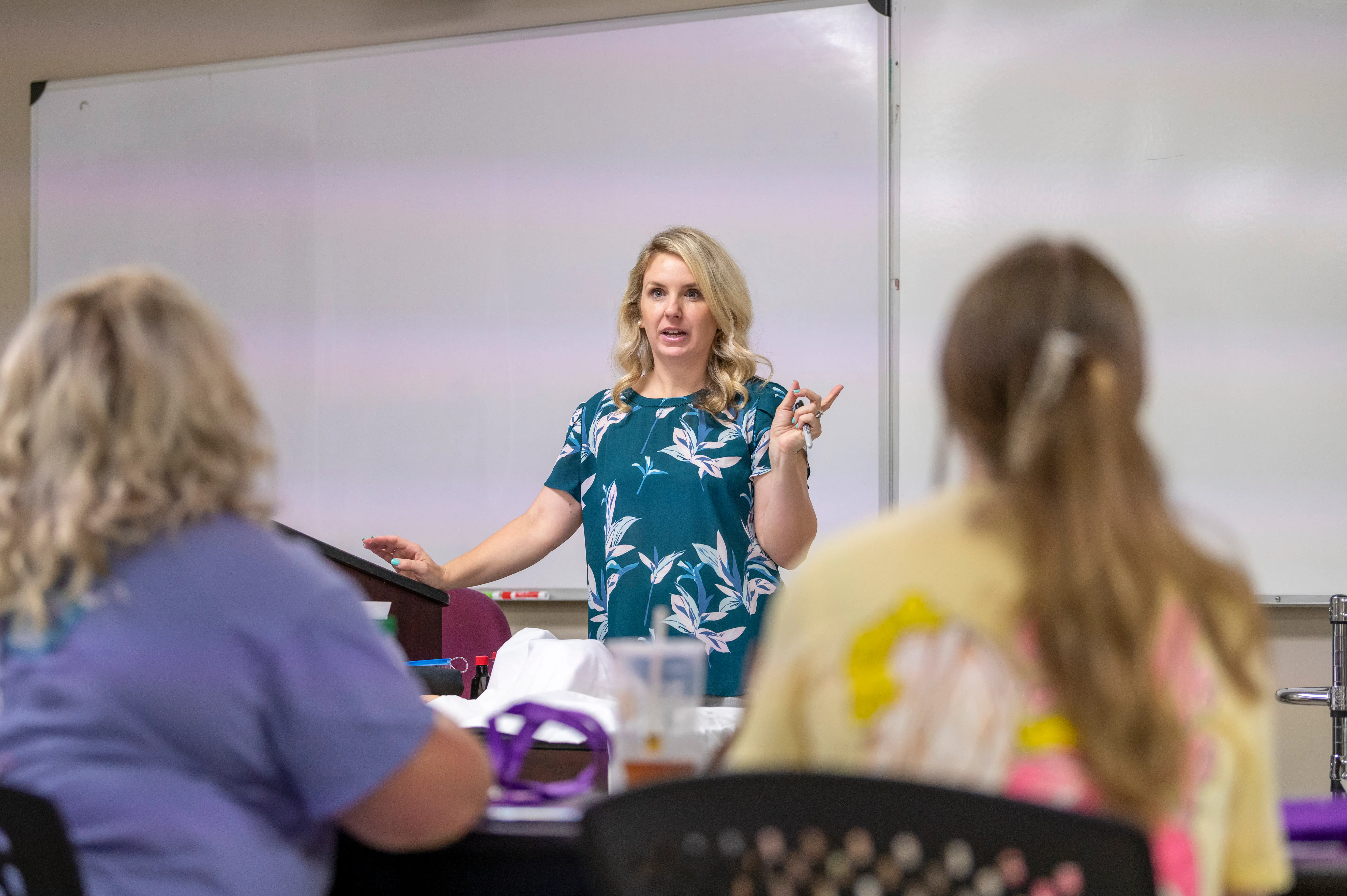 A nursing instructor addresses students during a classroom lecture, gesturing expressively in front of a whiteboard as two students look on from the foreground.