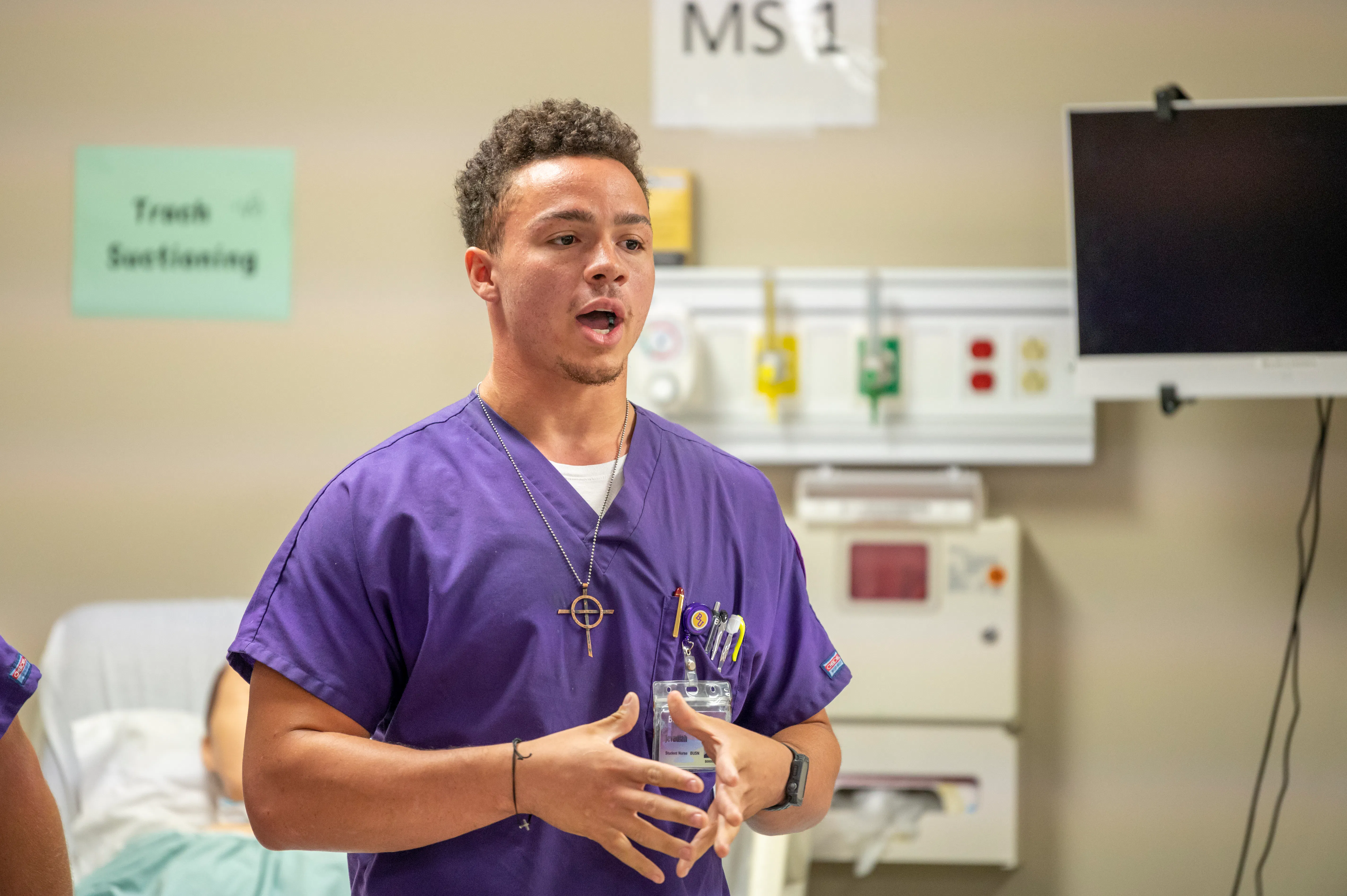 A nursing student in purple scrubs speaks during a clinical simulation lab exercise, with a patient mannequin and medical equipment visible in the background.