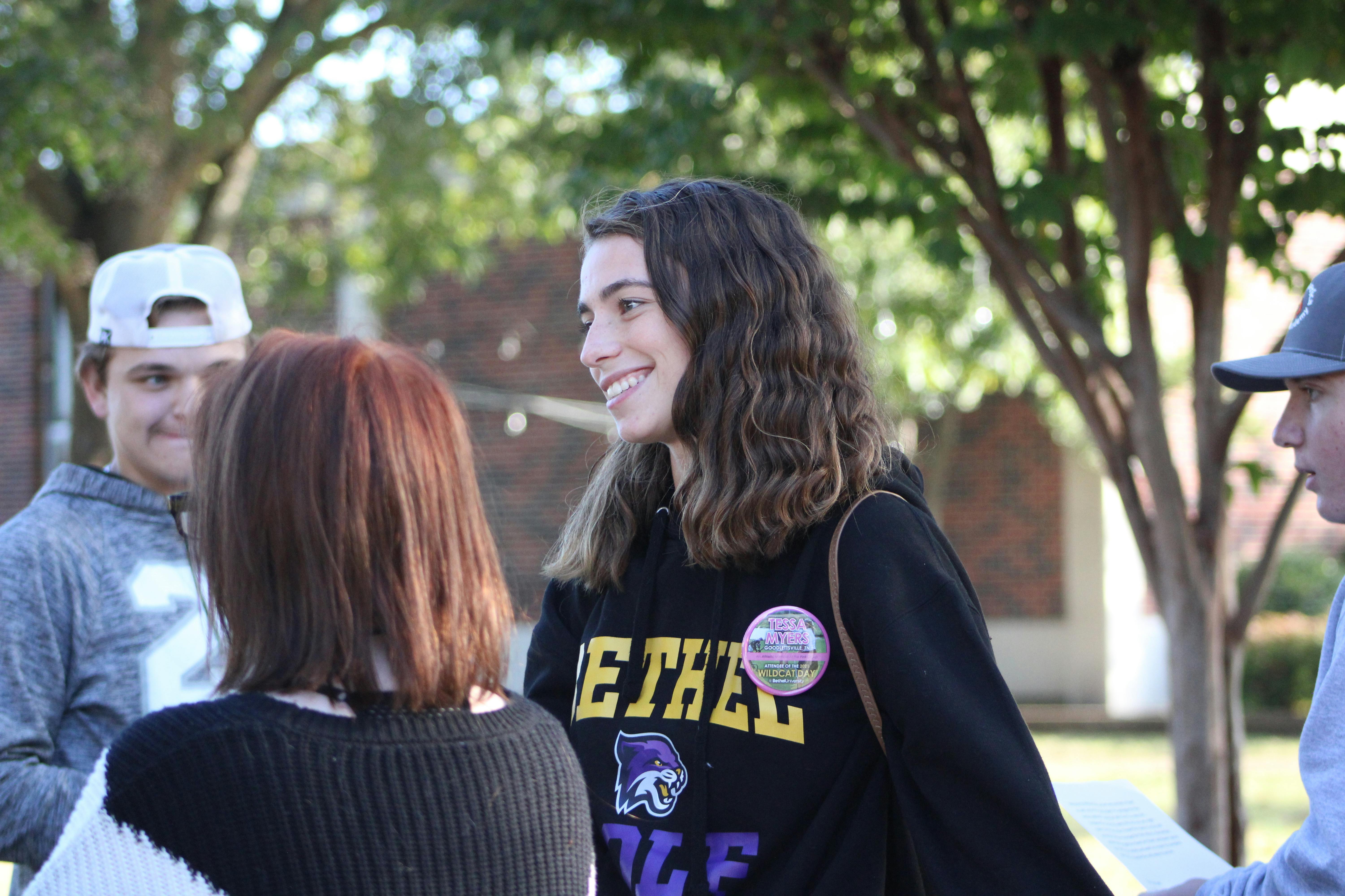 Student wearing a Bethel University sweatshirt smiles while talking with other students outdoors on campus.