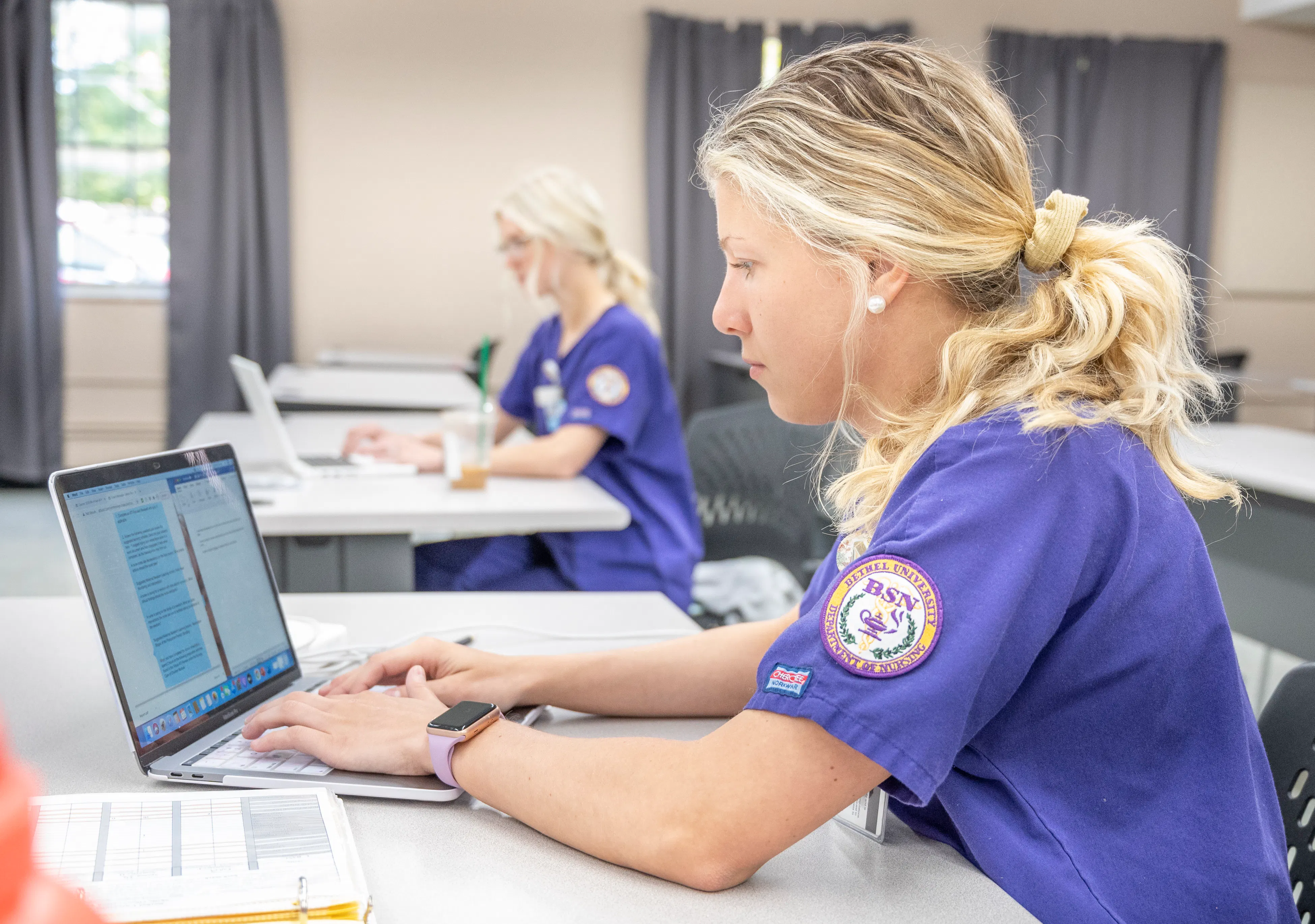 A Bethel University BSN nursing student works on a laptop in a classroom, with a second nursing student studying in the background.