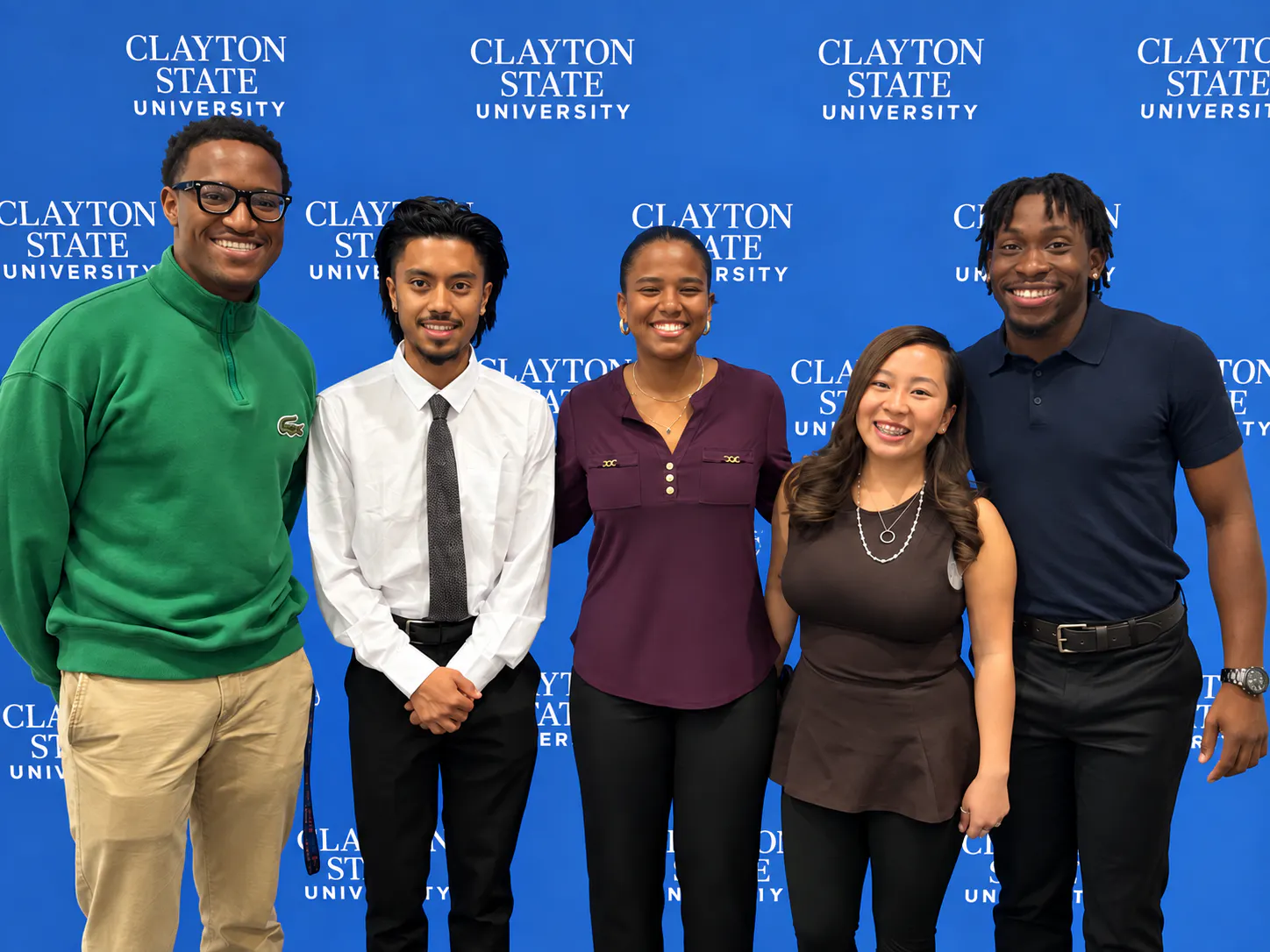 Five smiling college students standing side-by-side in front of a blue backdrop patterned with "Clayton State University" text.
