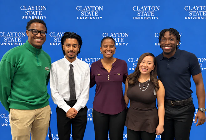 Five smiling college students standing side-by-side in front of a blue backdrop patterned with "Clayton State University" text.