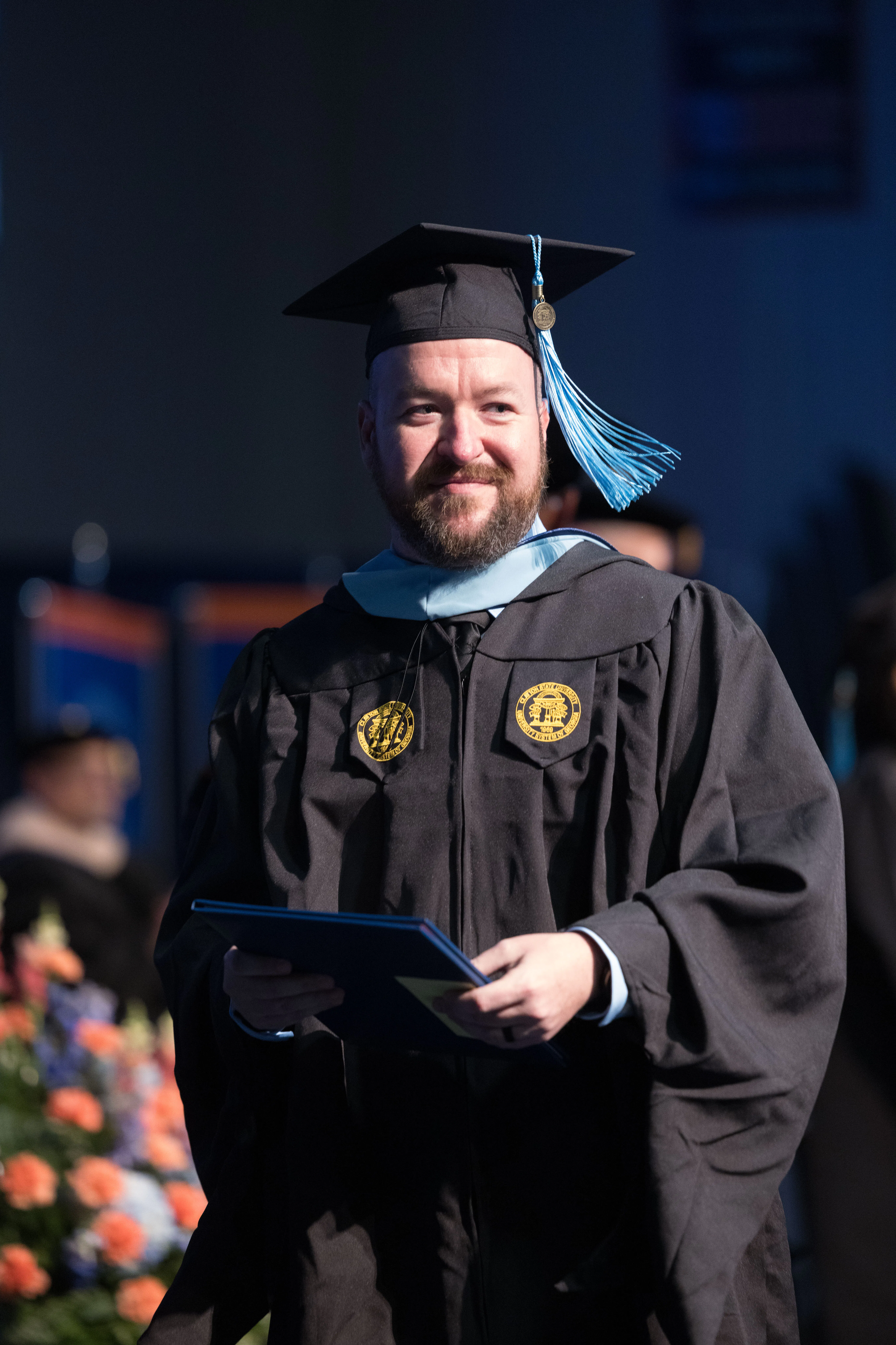 Smiling graduate in cap and gown holding a diploma onstage at commencement.