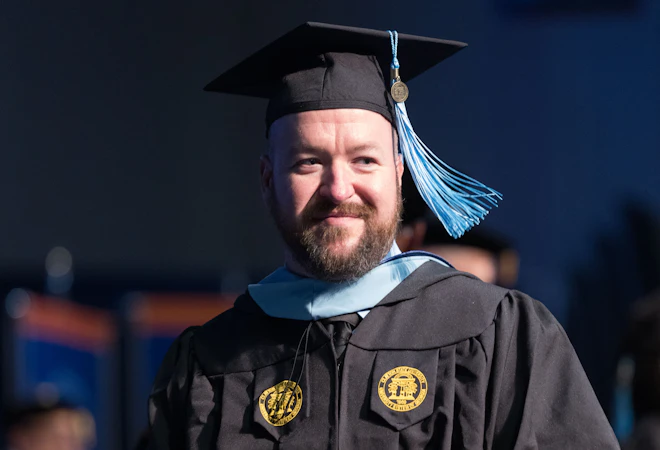Smiling graduate in cap and gown holding a diploma onstage at commencement.