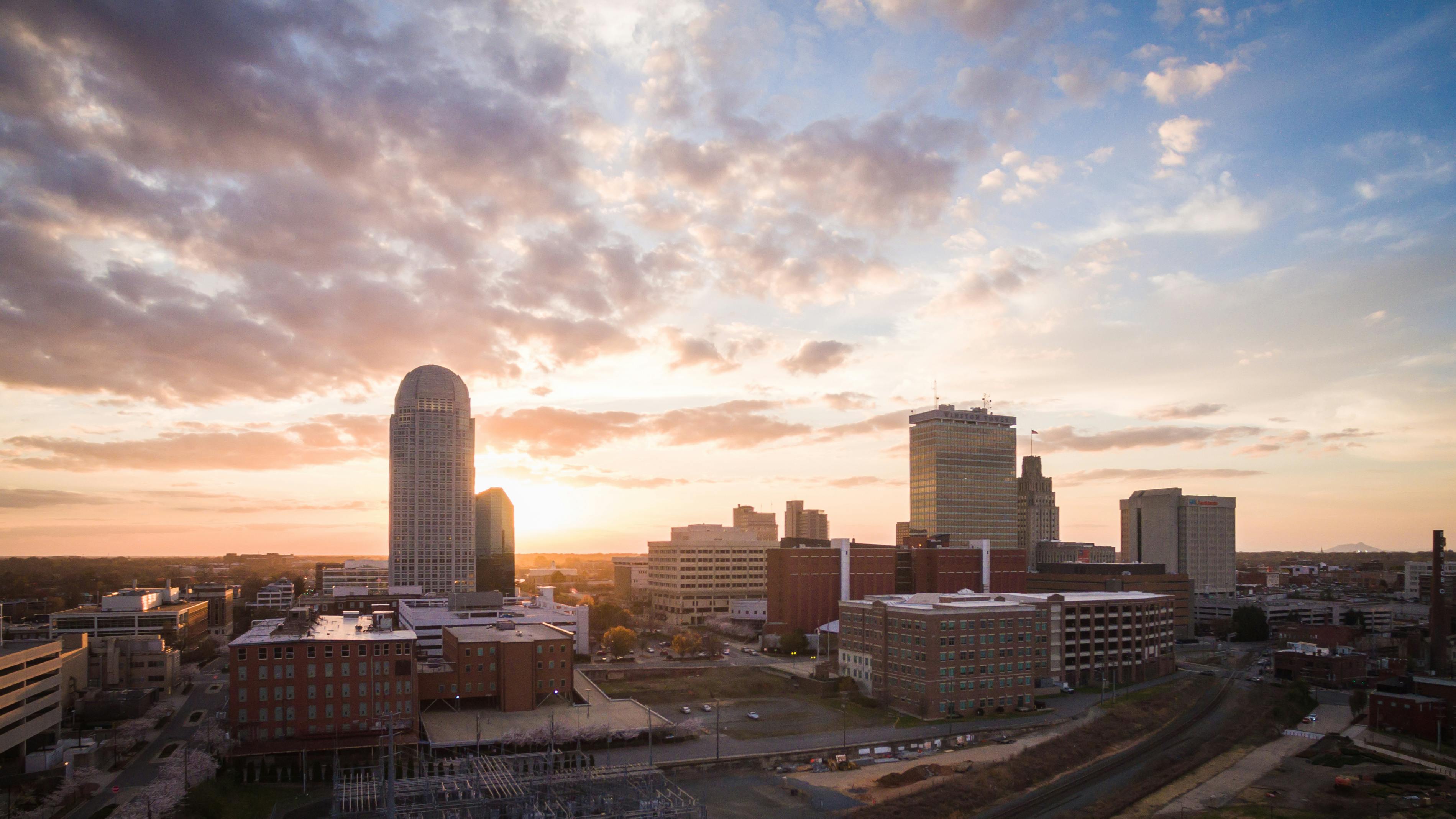 You've shared an image of a city skyline during what appears to be either dawn or dusk, as indicated by the soft, warm hues in the sky. The skyline is made up of several mid-rise and high-rise buildings, one of which has a distinctive cylindrical shape that stands out against the others. The sun appears low on the horizon, bathing the scene in orange and yellow light, which suggests either the beginning or end of the day.

The perspective is aerial, suggesting the photograph was taken from a height, such as a taller building or a drone. Below, we can see some urban infrastructure, including smaller buildings, parking lots, and streets that appear mostly devoid of traffic.