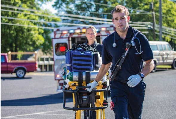 The image depicts two emergency medical service (EMS) responders in an outdoor environment during the day. The focal point is a young male EMS worker in the foreground, who appears attentive and is looking towards the camera. He is dressed in a standard EMS uniform, which includes a dark polo shirt with visible reflective stripes and EMS utility pants. He is wearing disposable gloves and has a stethoscope around his neck, signaling his readiness to provide medical assistance. He's pushing a wheeled stretcher or gurney that carries medical equipment, such as a blue medical bag likely containing essential medical supplies and a portable oxygen cylinder secured on the gurney.

Behind him, slightly out of focus, is another EMS worker who appears to be looking on or supervising the situation. An ambulance with its back doors open is visible in the background, indicating that they are either arriving at a scene or preparing to transport a patient. The environment suggests a suburban setting, with trees, clear skies, and power lines overhead.