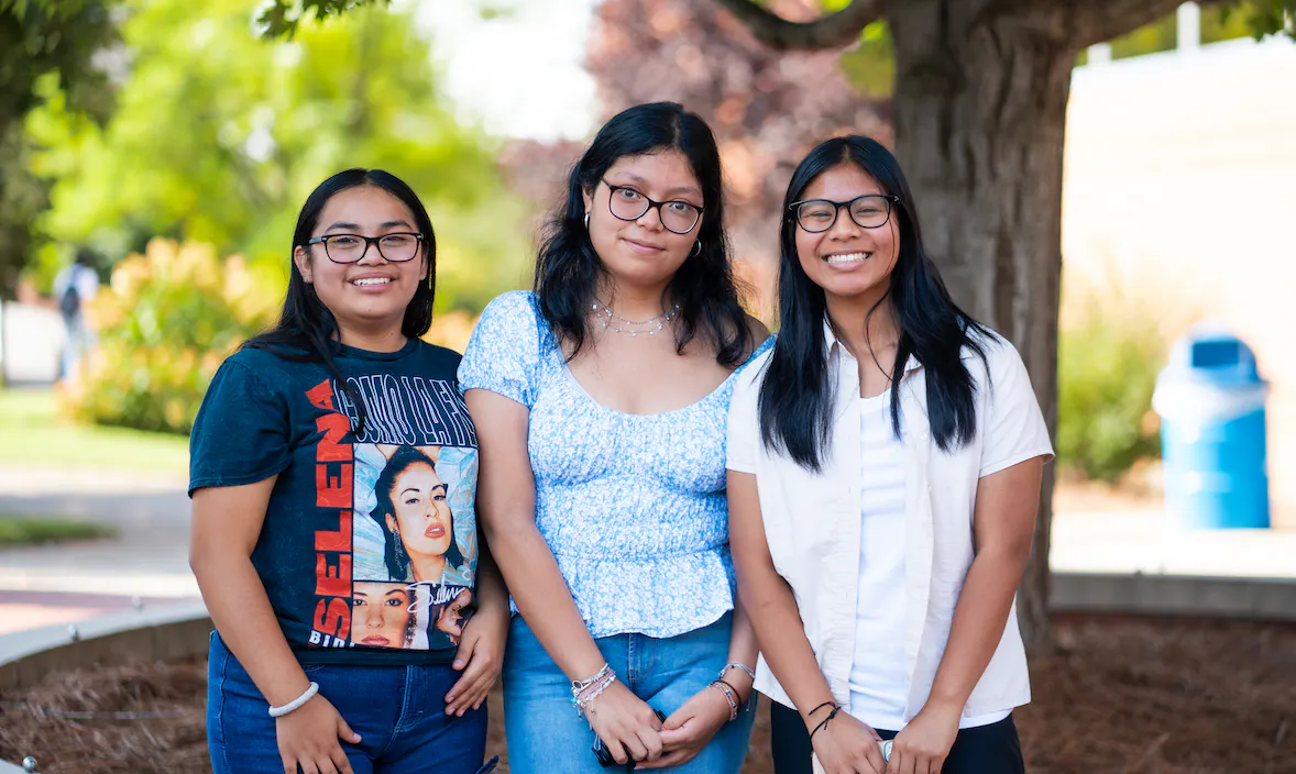 The image features three young women posing together outdoors. They are standing in what appears to be a campus setting with greenery and a tree in the background, suggesting a college or university environment. The woman on the left is wearing glasses, a graphic t-shirt with red accents, and jeans. The woman in the center is wearing a blue patterned top and blue jeans, and the woman on the right is dressed in a white button-up shirt and black pants. All three are wearing eyeglasses and have their hair down.