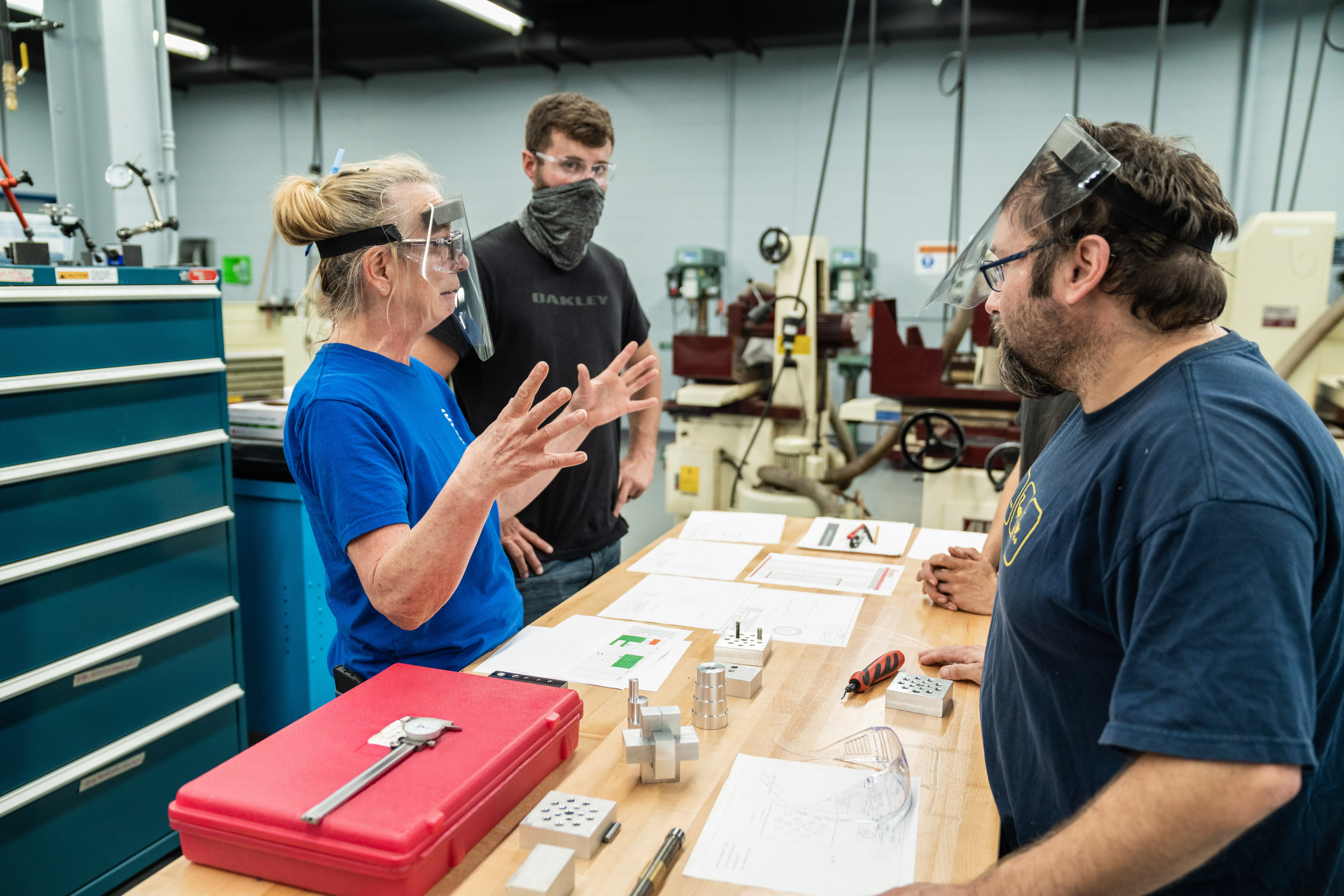 The image captures a scene in what appears to be a vocational training or educational workshop setting. Three individuals are engaged in a discussion or instructional activity. The person on the left is a woman wearing a blue shirt, protective goggles, and a face shield, and she is gesturing with her hands, possibly explaining a concept or giving instructions. The person in the middle is a younger male, wearing a black shirt and a protective mask covering his mouth and nose, observing the interaction. He is also wearing safety goggles. On the right, there's a man with a beard, wearing a blue shirt similar to the woman's, goggles resting on his forehead, and he is turning towards the woman, likely listening or responding to her.

In front of the individuals is a workbench with various tools and educational materials, including paper documents, which could be instructional guides or project plans. They appear to be in a learning environment with a focus on hands-on skills or trades education. The background shows additional workshop equipment and workspaces, suggesting a practical educational setting, possibly in a higher education institution like a community college or technical institute.