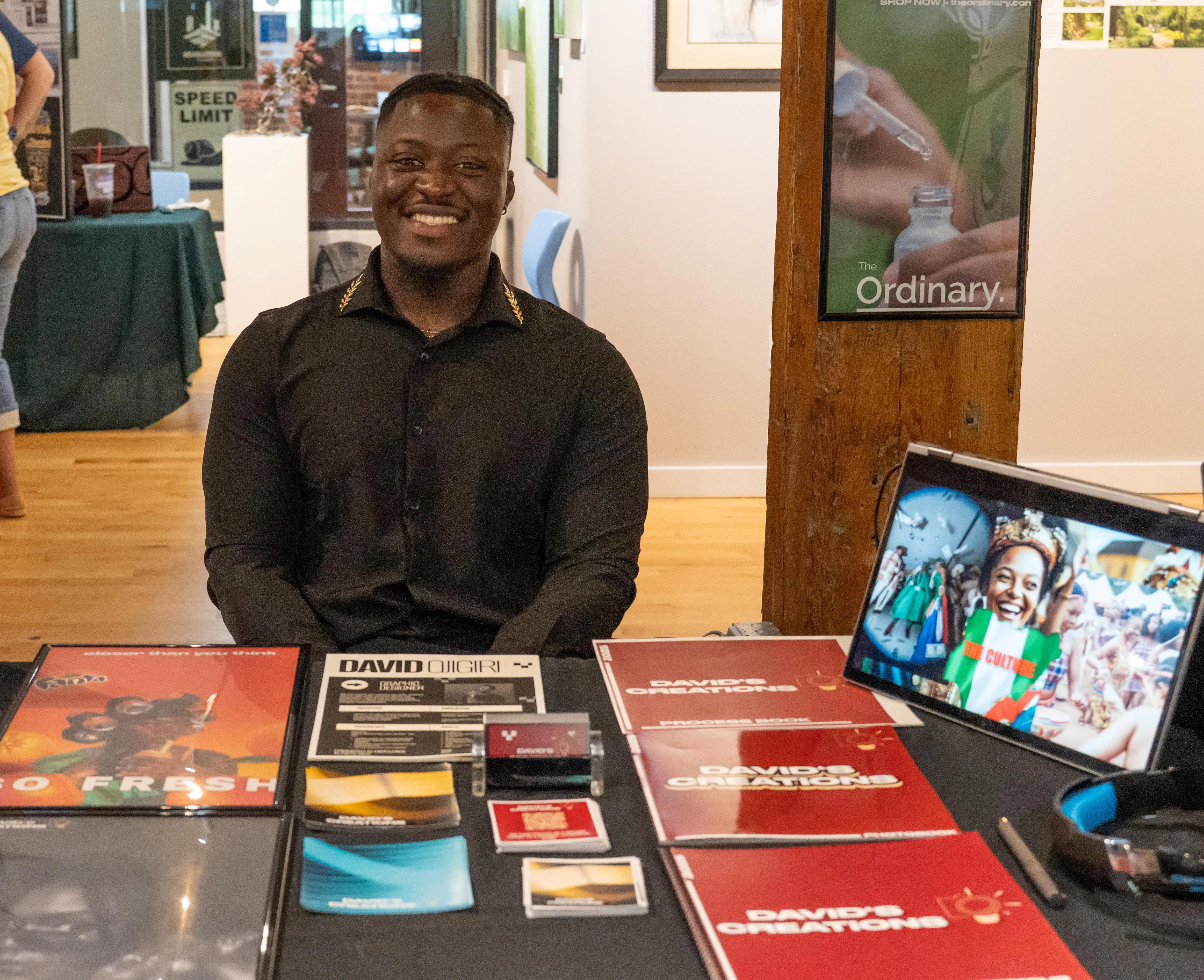 A smiling student sits behind a display table showcasing design and photography work, including printed portfolios and business cards. A tablet on the table displays a digital project, and posters and displays are visible in the background, suggesting a student showcase or portfolio event.