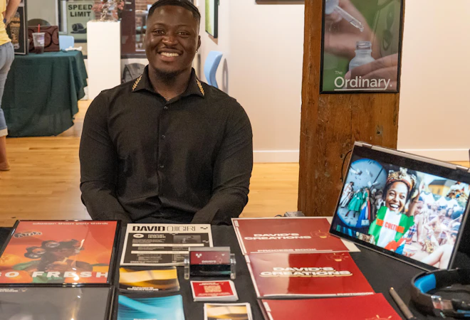 A smiling student sits behind a display table showcasing design and photography work, including printed portfolios and business cards. A tablet on the table displays a digital project, and posters and displays are visible in the background, suggesting a student showcase or portfolio event.