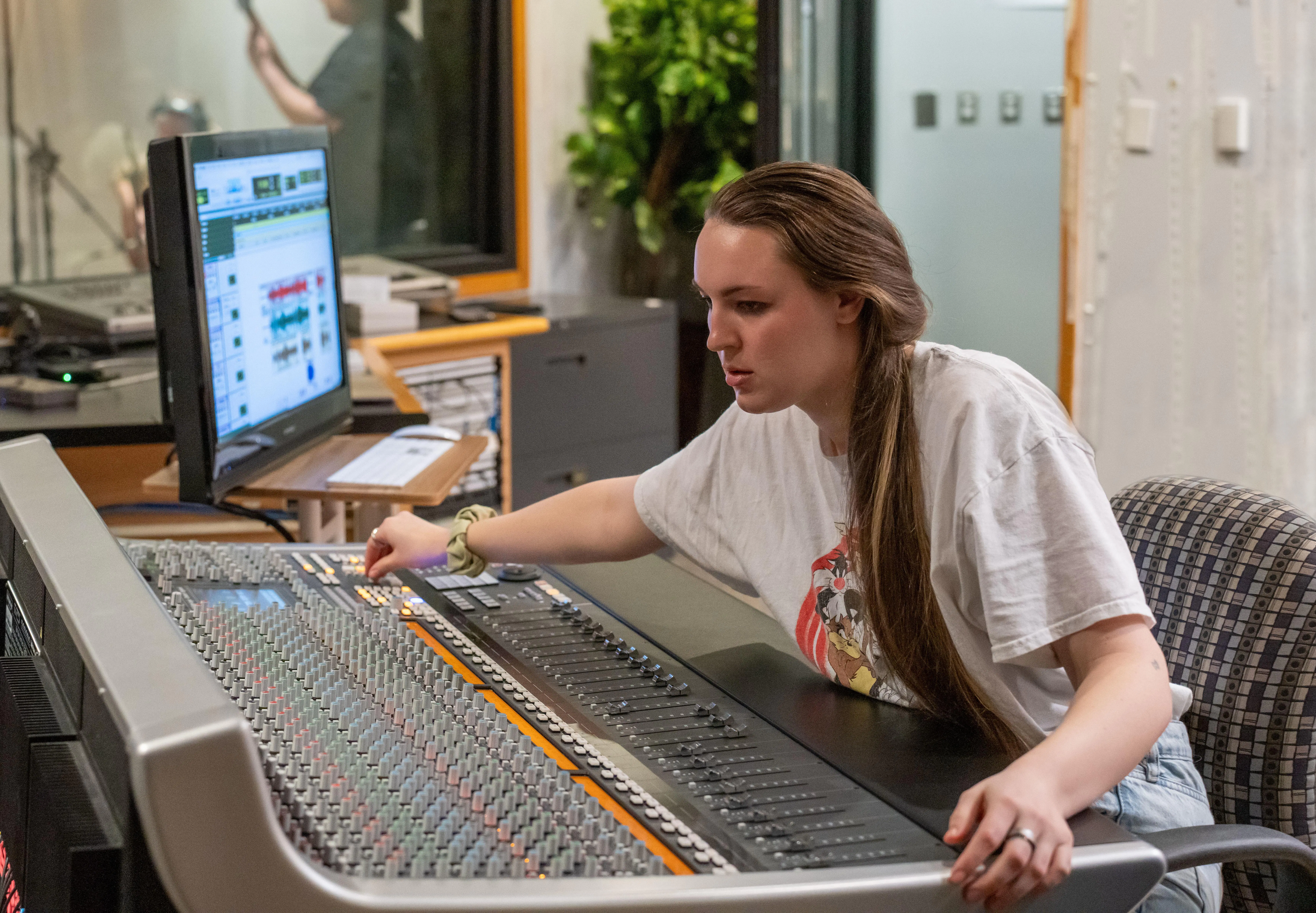 A student sits at a large audio mixing console in a recording studio, adjusting controls while focusing on the sound levels displayed on a nearby computer monitor. A microphone booth is visible in the background, suggesting an active recording session.