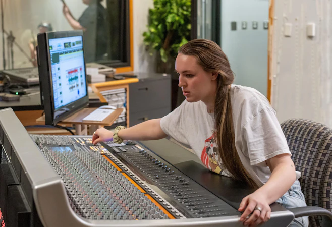 A student sits at a large audio mixing console in a recording studio, adjusting controls while focusing on the sound levels displayed on a nearby computer monitor. A microphone booth is visible in the background, suggesting an active recording session.