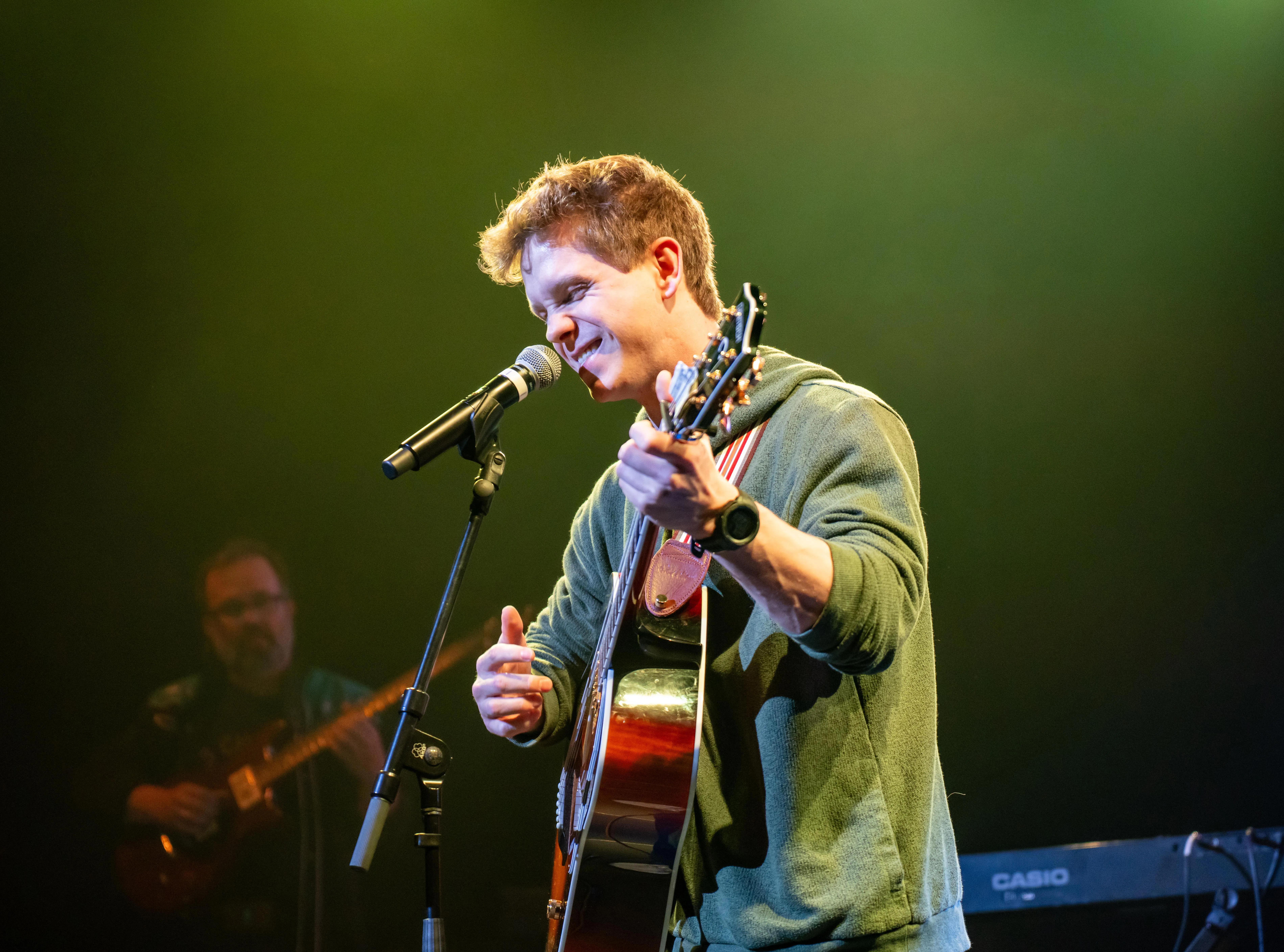 A musician performs on stage, singing into a microphone while playing an acoustic guitar. Colored stage lighting illuminates the scene, and another band member is visible in the background.