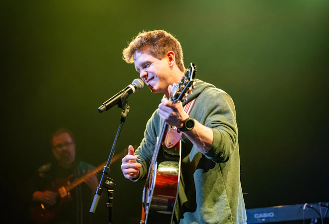 A musician performs on stage, singing into a microphone while playing an acoustic guitar. Colored stage lighting illuminates the scene, and another band member is visible in the background.