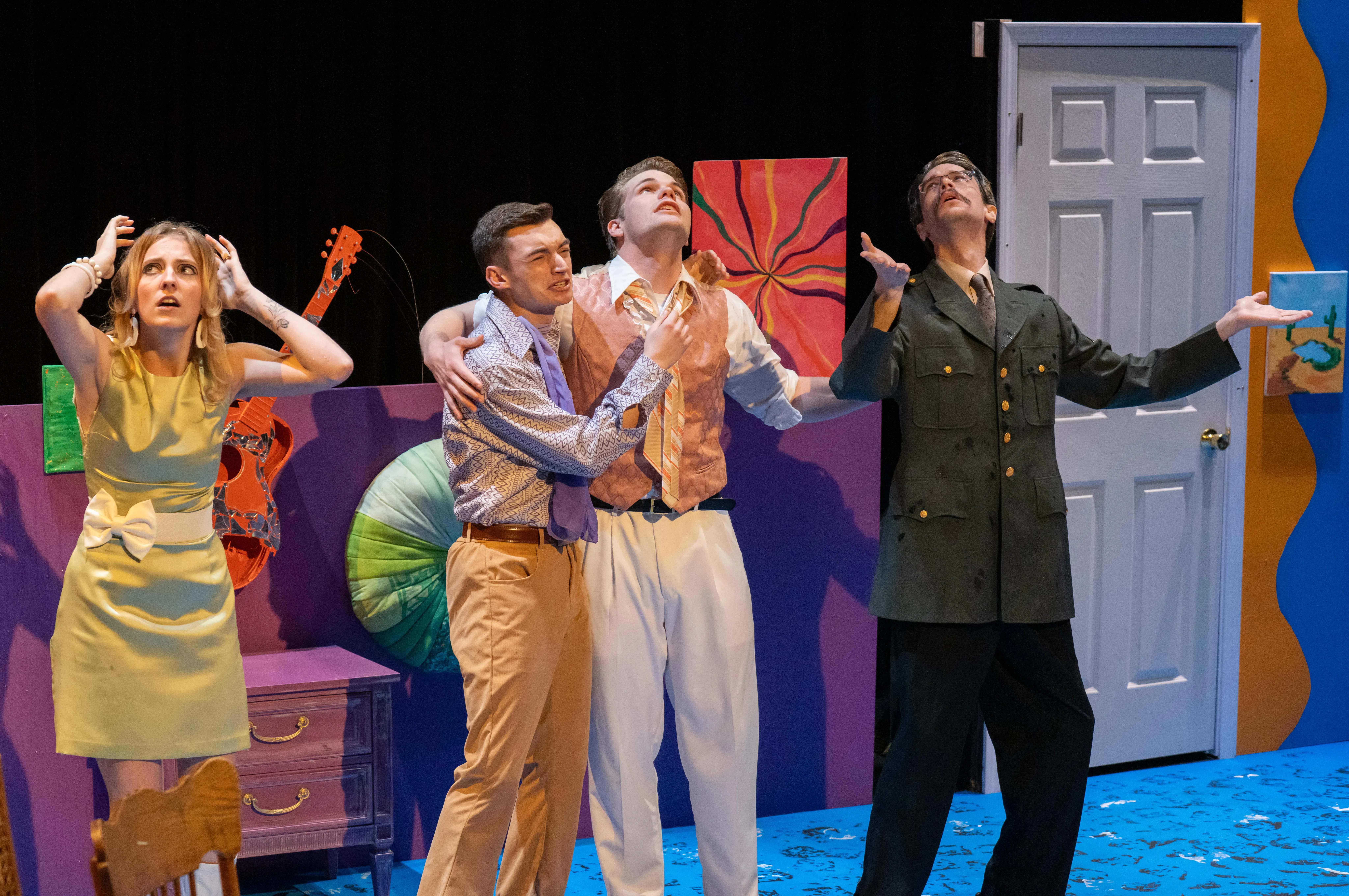 Four actors perform on a colorful stage set, expressing exaggerated emotions during a theatrical scene. One character holds another dramatically, while a third gestures upward in a military-style costume, and a fourth reacts with hands on their head, suggesting a moment of heightened tension or comedy.