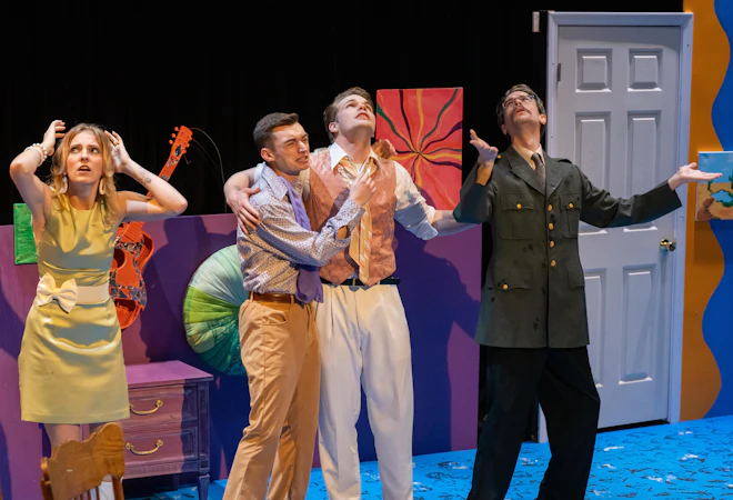 Four actors perform on a colorful stage set, expressing exaggerated emotions during a theatrical scene. One character holds another dramatically, while a third gestures upward in a military-style costume, and a fourth reacts with hands on their head, suggesting a moment of heightened tension or comedy.