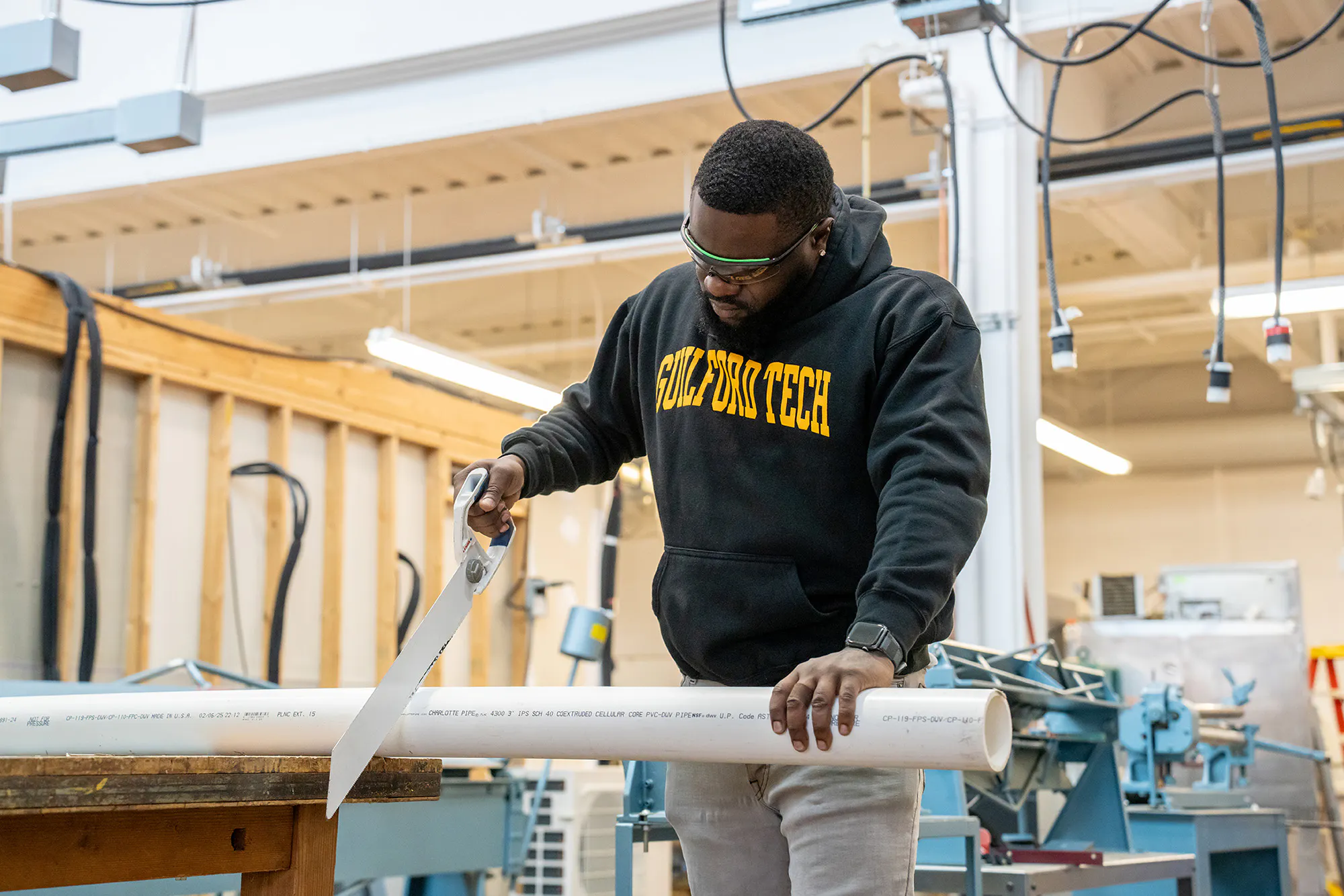 Male plumbing student sawing pipe. He is wearing a black Guilford Tech sweatshirt with gold lettering.