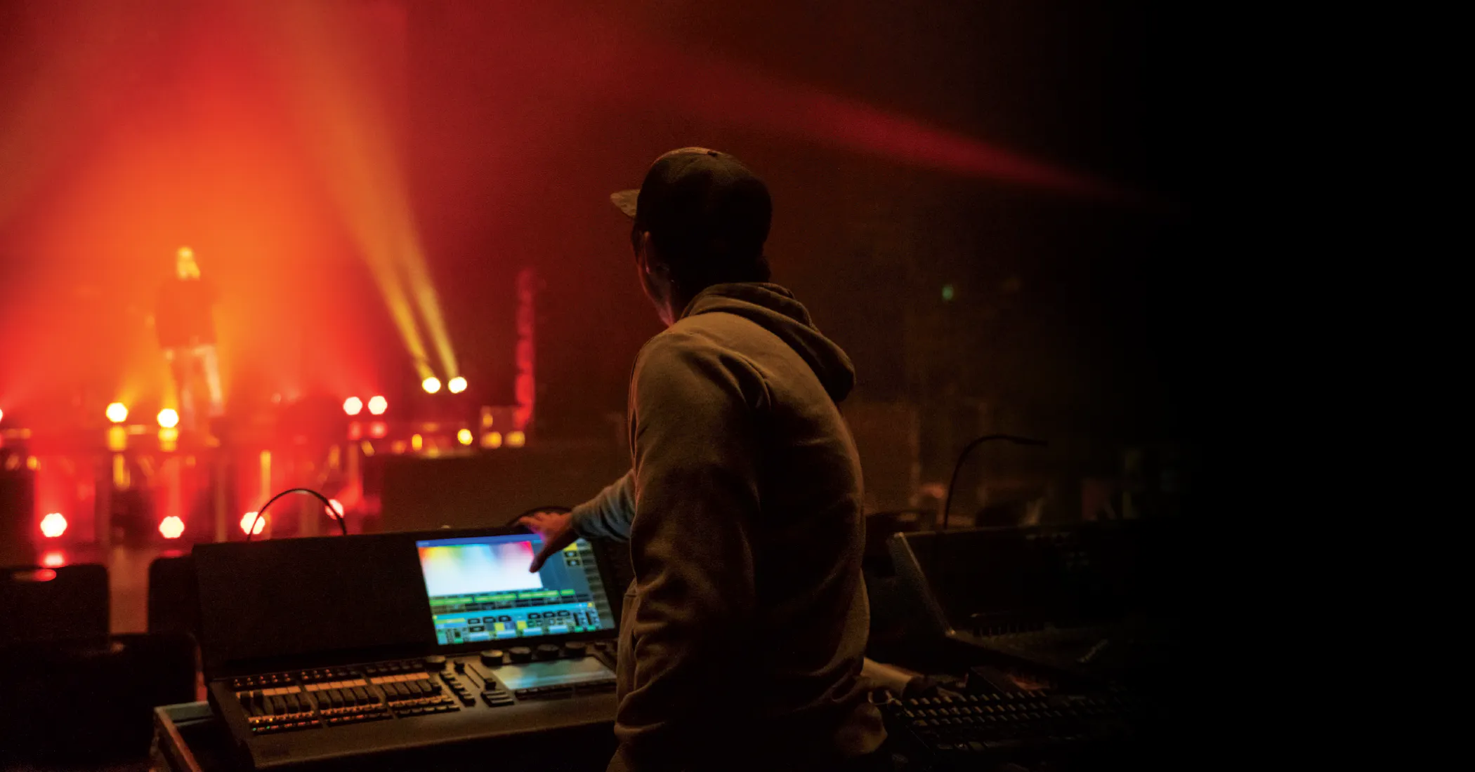 A student operates a lighting or sound control console in a dark theater, adjusting settings while looking toward a stage illuminated with colorful lights. A performer stands in the distance, suggesting a live production or rehearsal in progress.