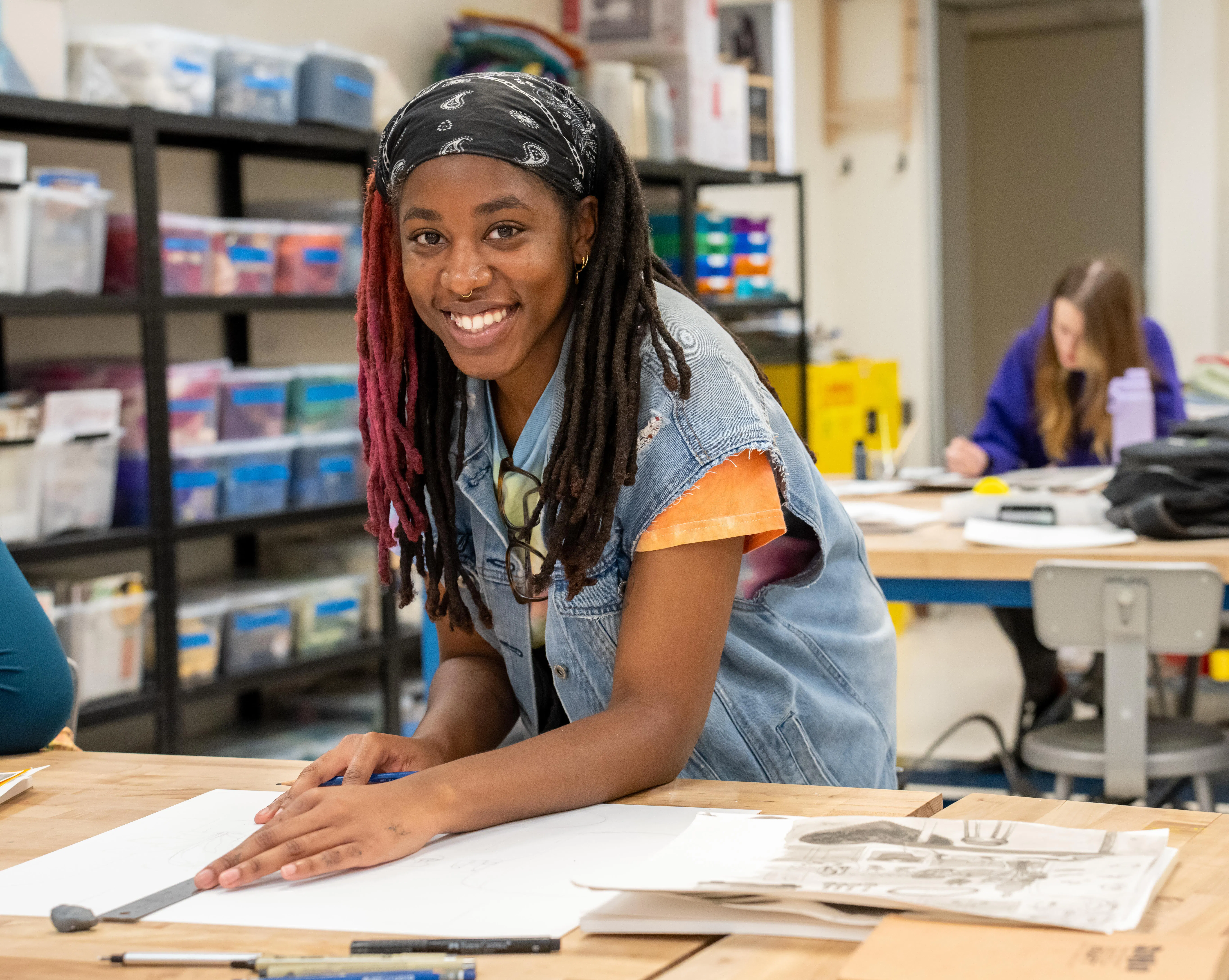 A student smiles at the camera while working on an art project at a classroom table, surrounded by drawing materials and papers. Shelves filled with supplies are visible in the background, and another student works independently at a nearby desk.