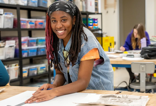 A student smiles at the camera while working on an art project at a classroom table, surrounded by drawing materials and papers. Shelves filled with supplies are visible in the background, and another student works independently at a nearby desk.