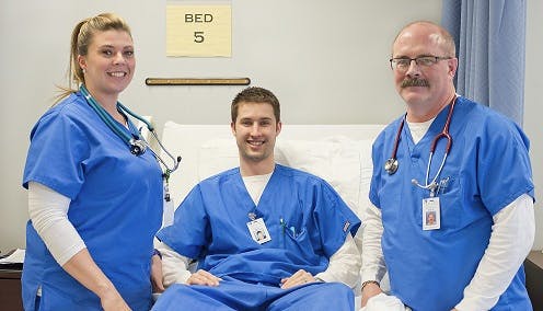 Nursing student administering an IV to a classmate while an instructor looks on