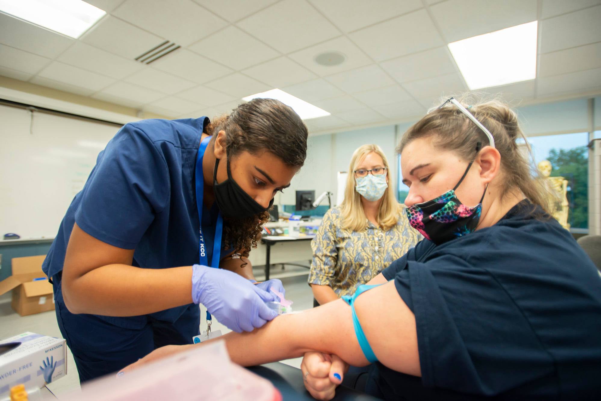 Nursing student administering an IV to a classmate while an instructor looks on