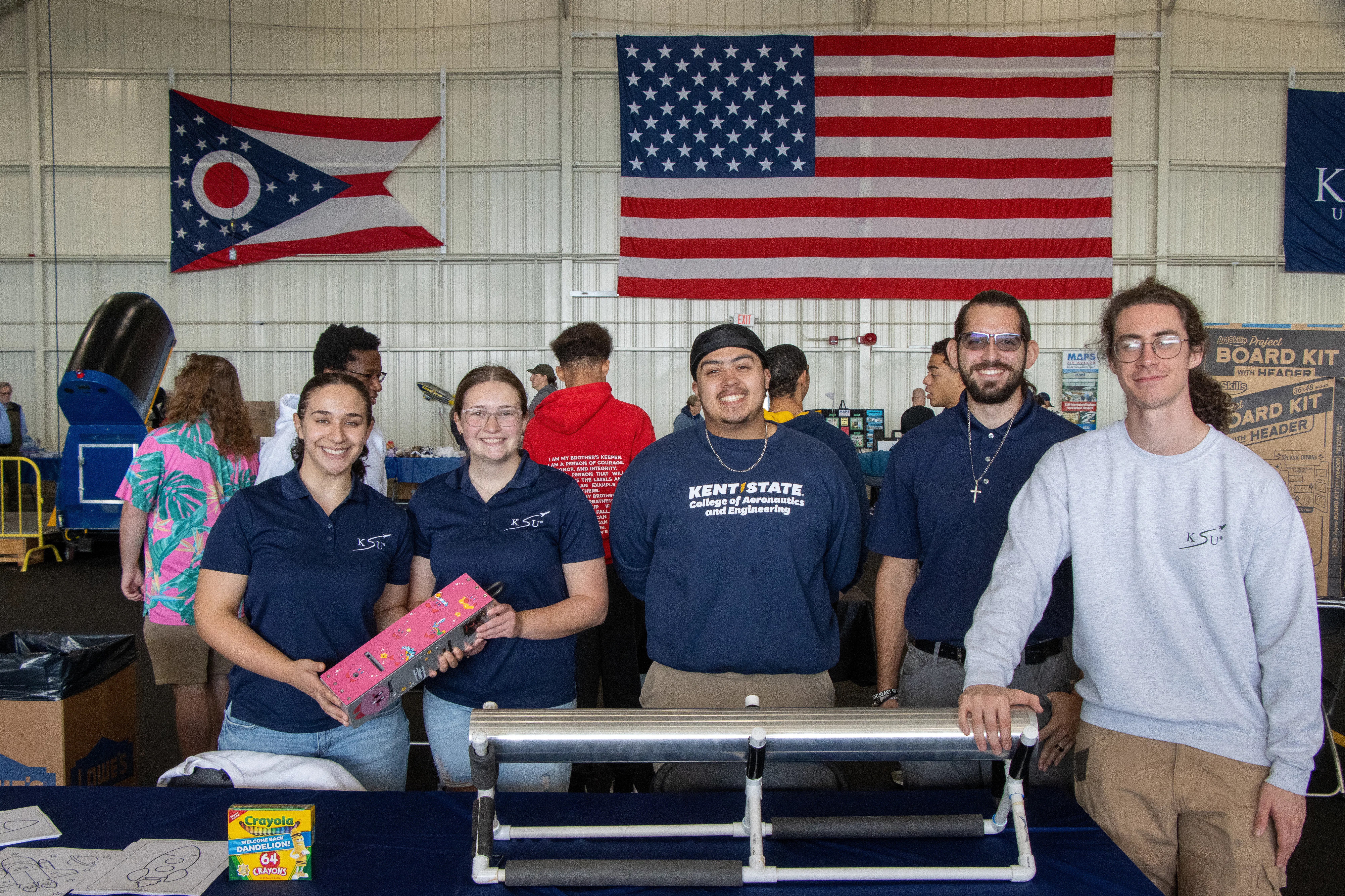 Two female and three male students in the hangar with an American flag in the background. The two femailes are wearing polo shirts with the KSU Rocket team logo. the male int he middle is wearing aKent State College of Aeronautics and Engineering branded dark blue crew neck. The next male is wearing a navy blue polo shirt, and the last one is wearing a heather gray crewneck sweatshirt. 