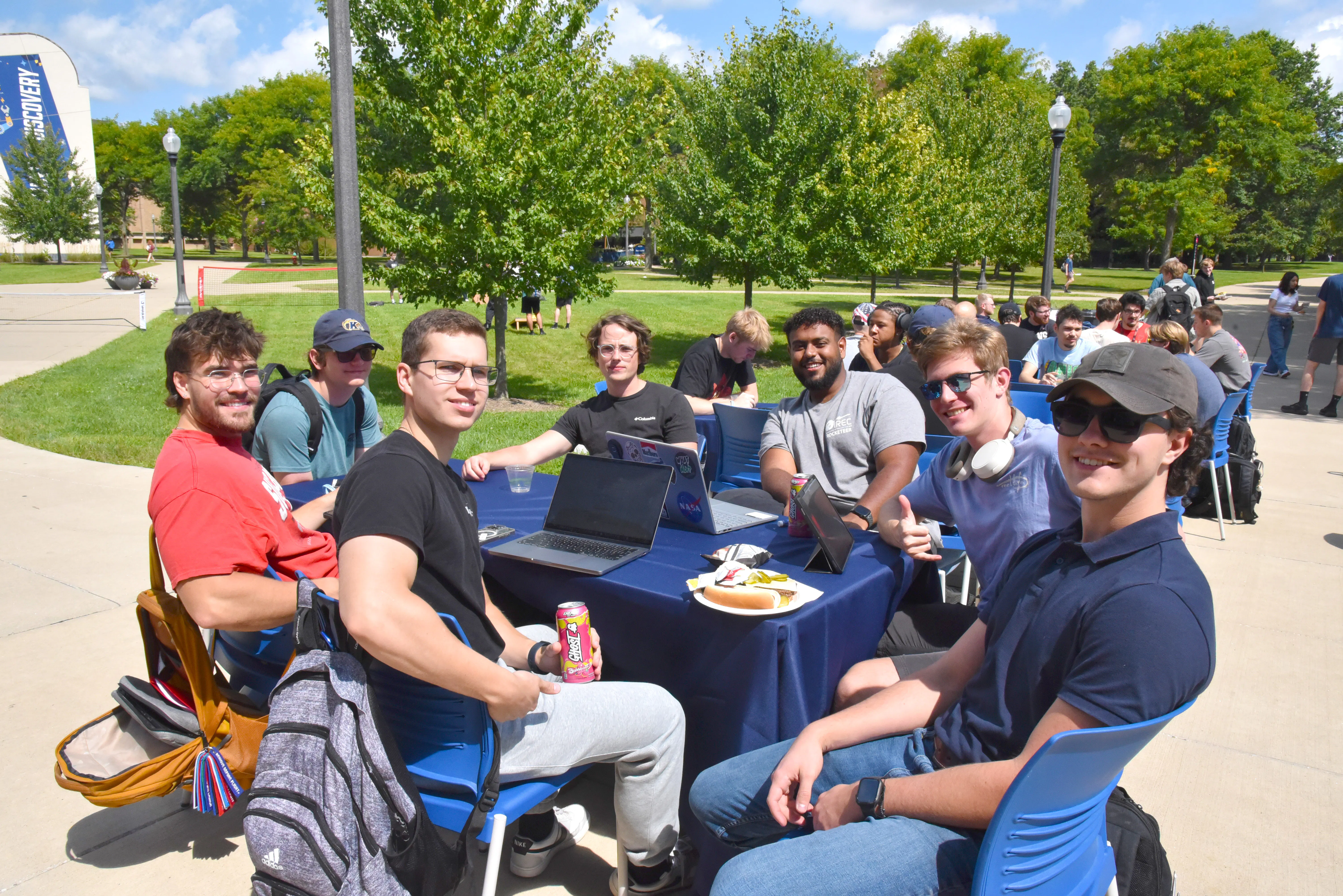 A group of seven male CAE students sit at a table outside to eat and socialize at the Kickoff Cookout, part of the Week of Welcome at the College of Aeronautics and Engineering. 