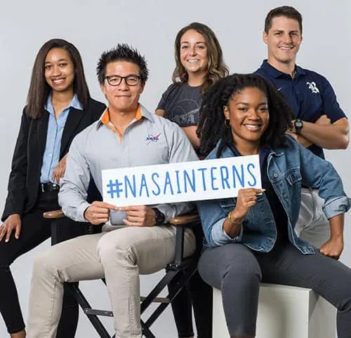 Diverse group of young adults dressed in business casual attire holding a sign that says #NASAInterns.