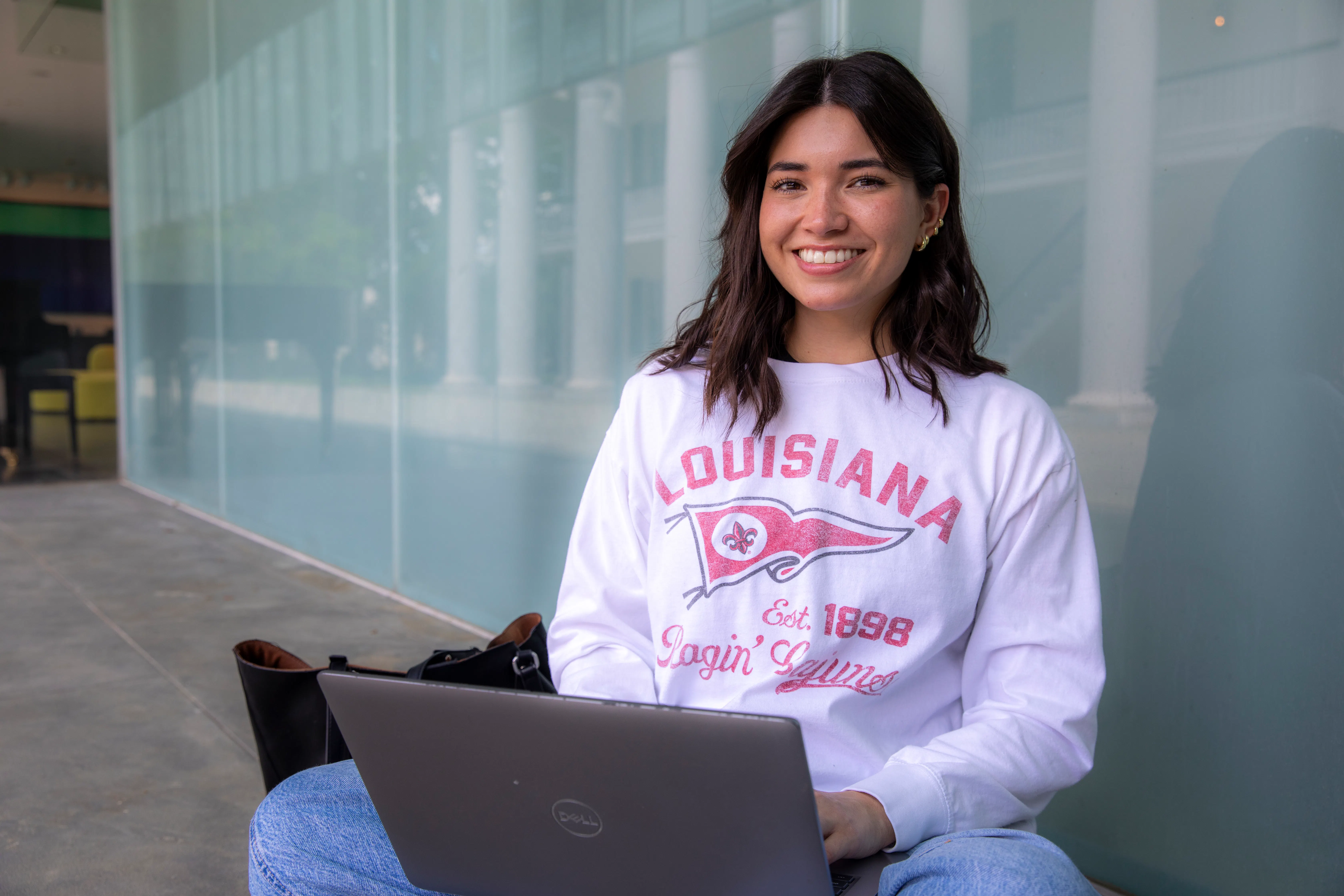 Online student sits crisscrossed while working on laptop and wearing a white, long-sleeve Ragin' Cajuns t-shirt.