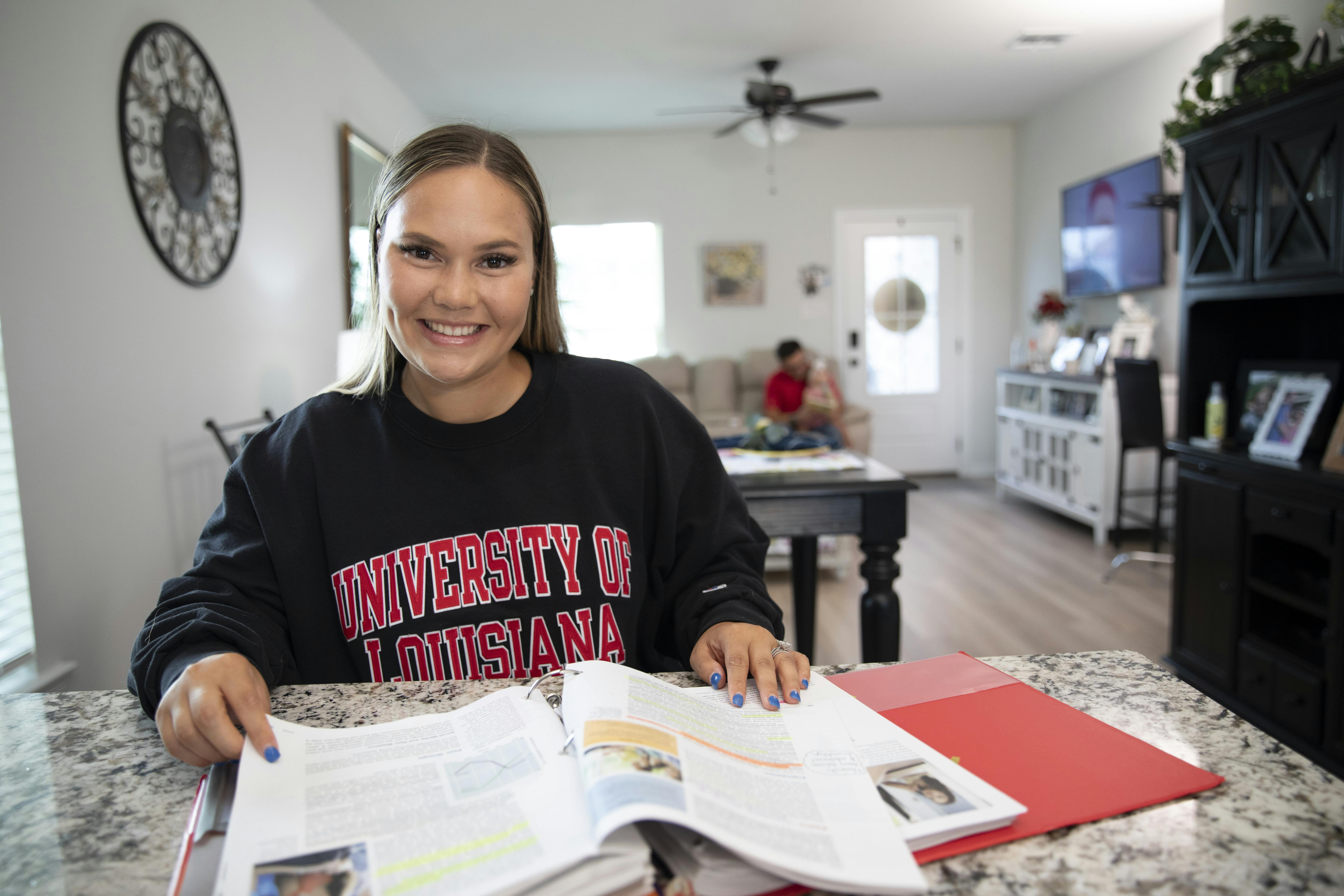 A woman is pictured in a University of Louisiana sweatshirt with a textbook in the foreground and her family in the background.