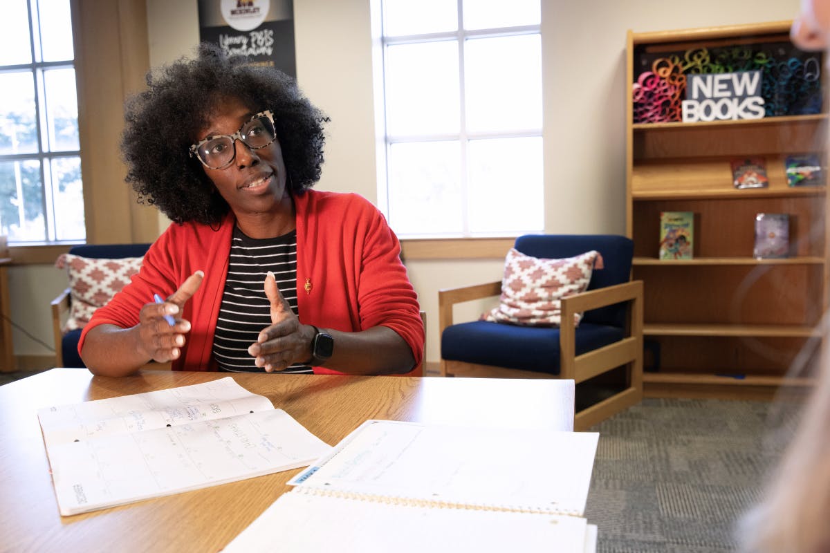 A woman is pictured in a red sweater as a school classroom work table.