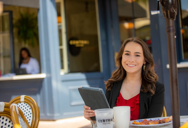 Health Services Administration alum Amanda Terry sits outdoors at a cafe table holding a tablet.