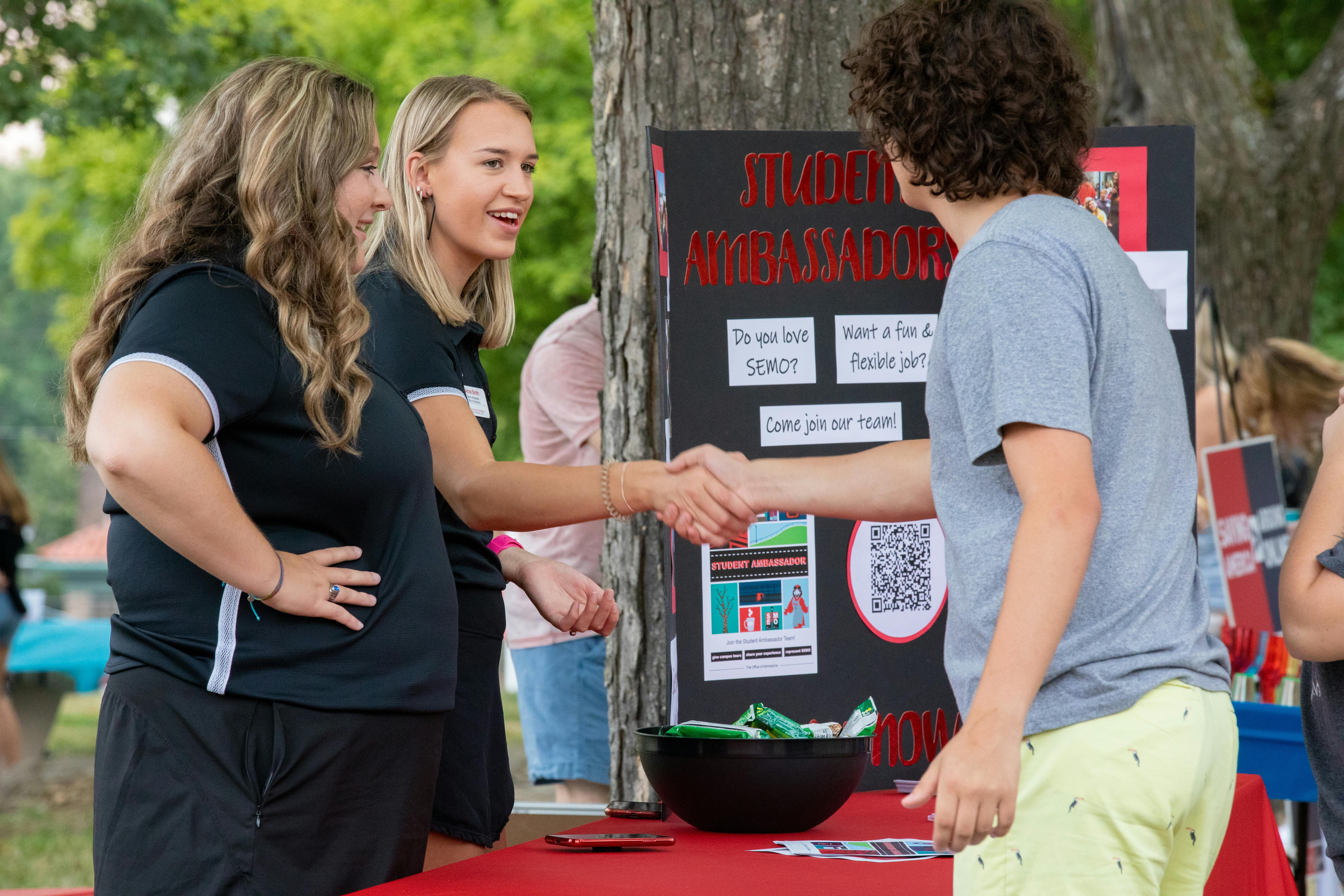 a prospective student greets a two students at an informational booth about student ambassadors. 