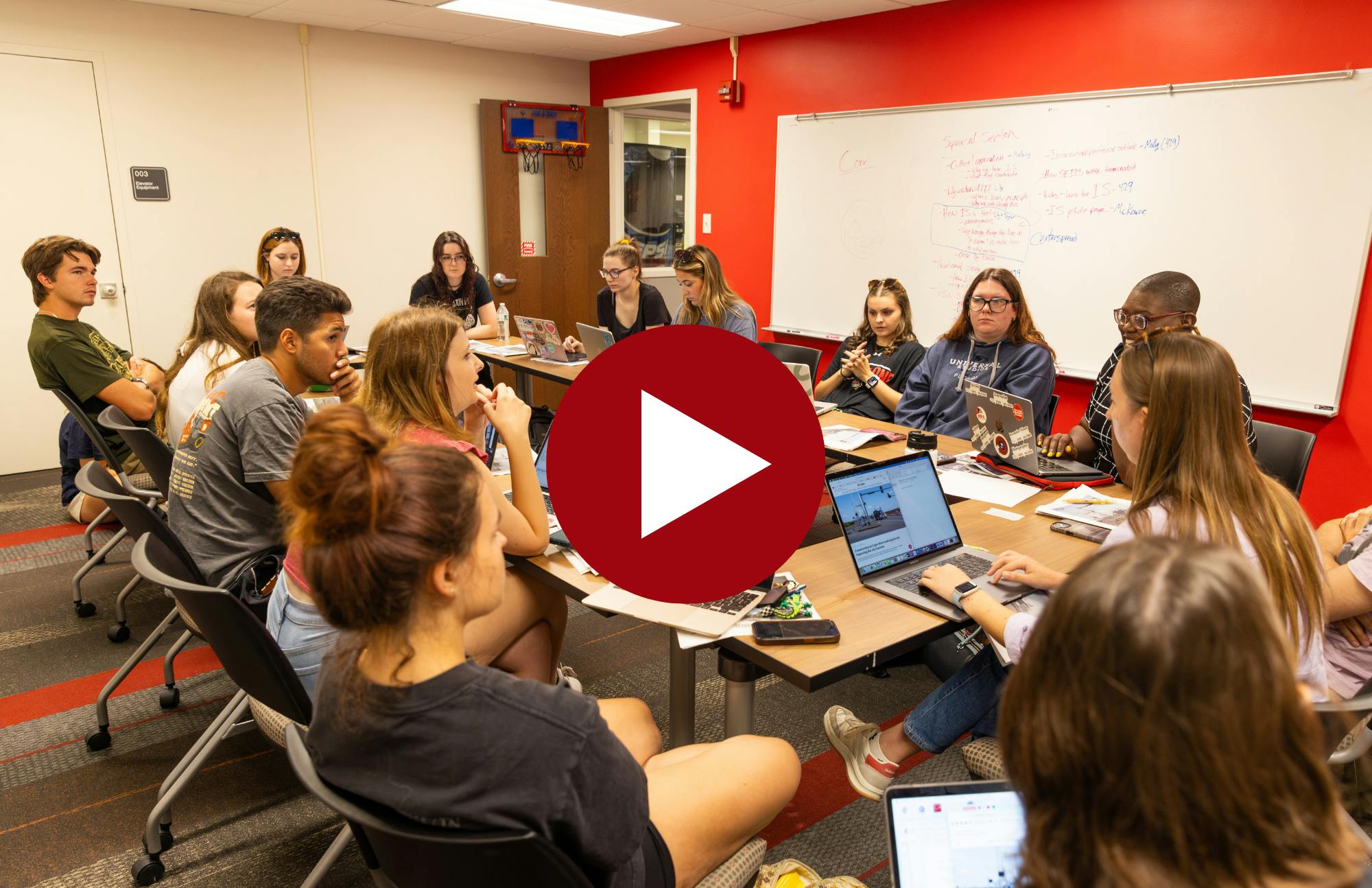 red play button over top image of mass media students gathered around tables in a "u" shape discussing content