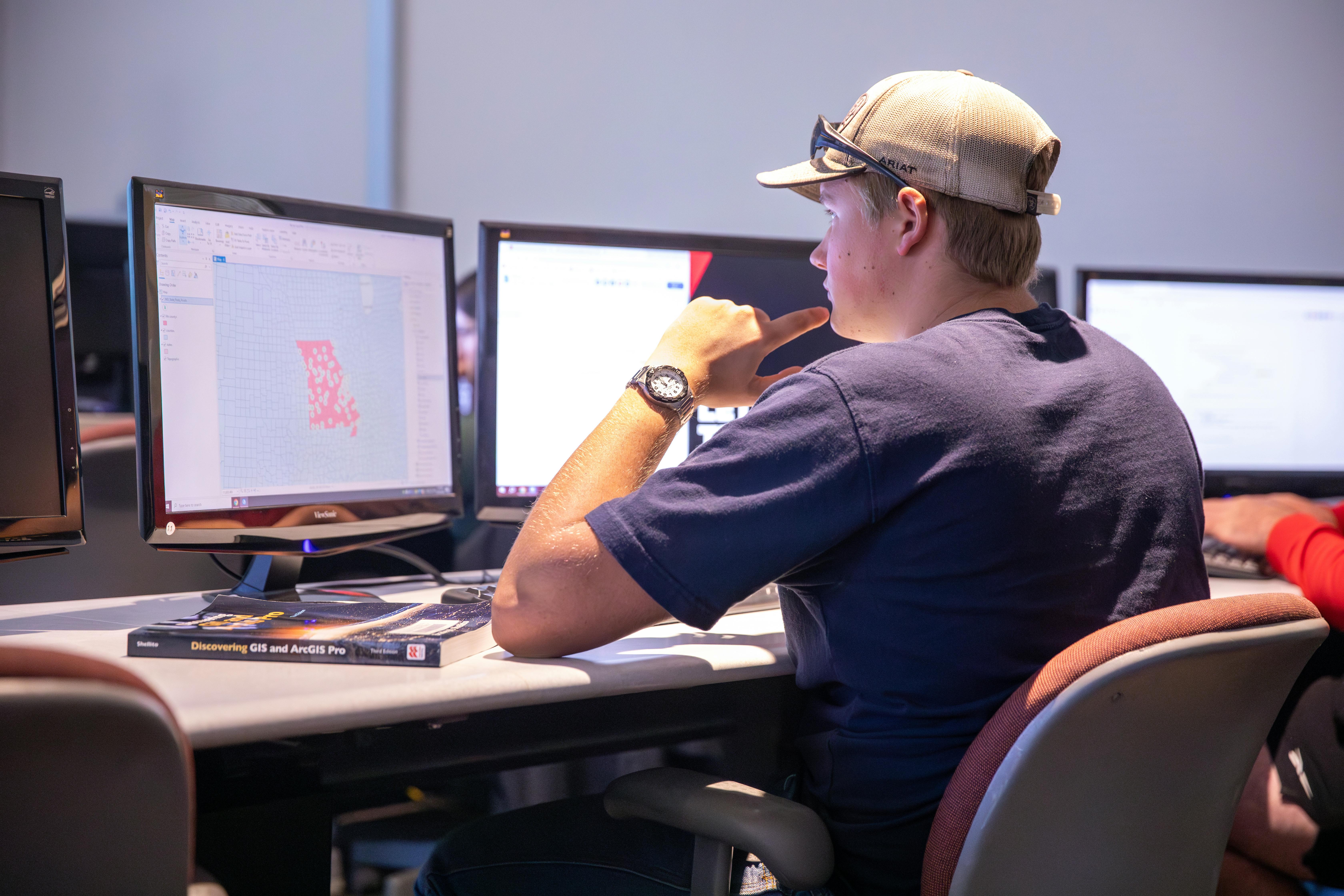 Man sits at desk with one finger resting on his chin looking at two computer screens. the screen on his left has a color-coded map of Missouri open. 