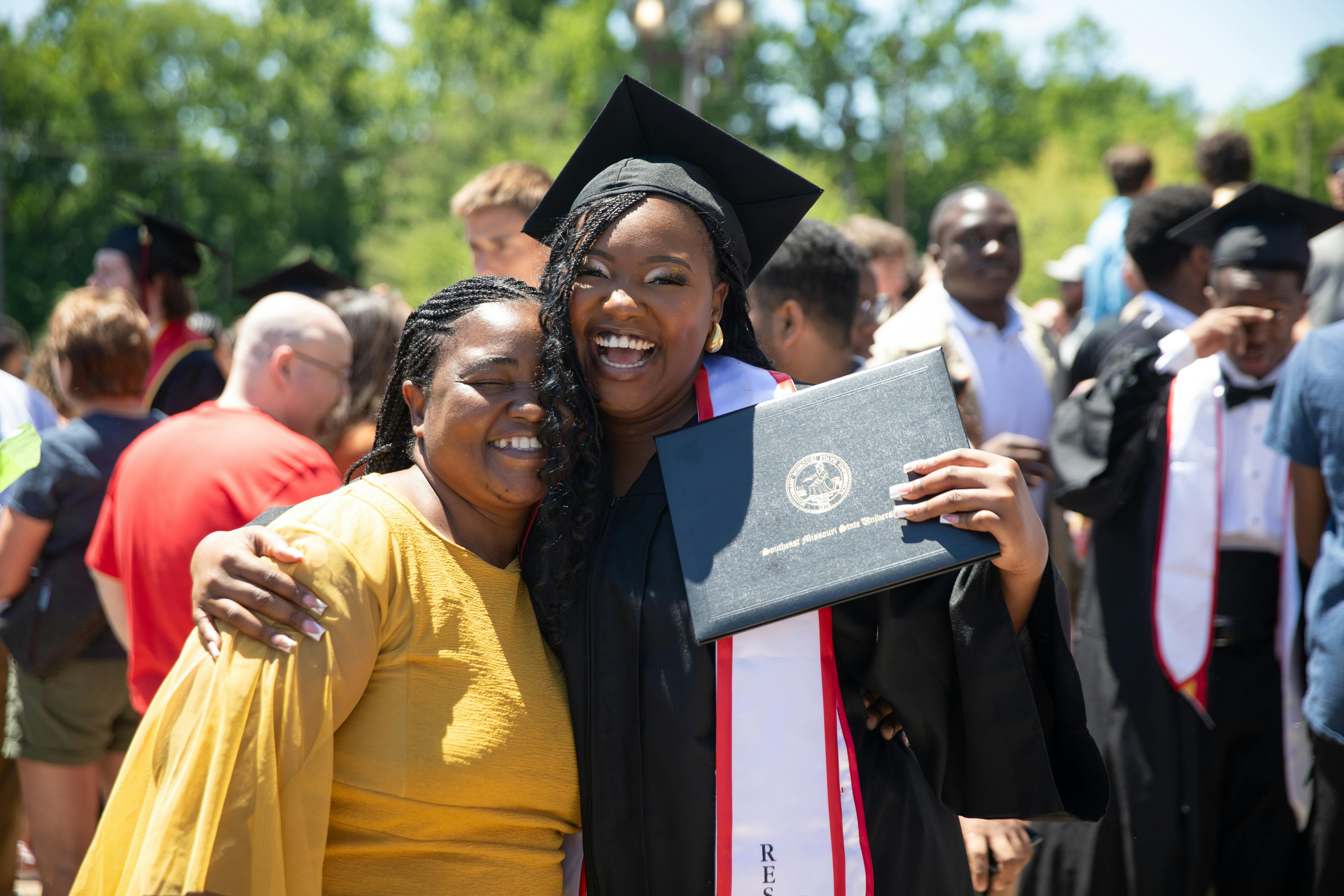 woman stands in her cap and gown outside of graduation holding up her diploma and hugging her mother from the size. 